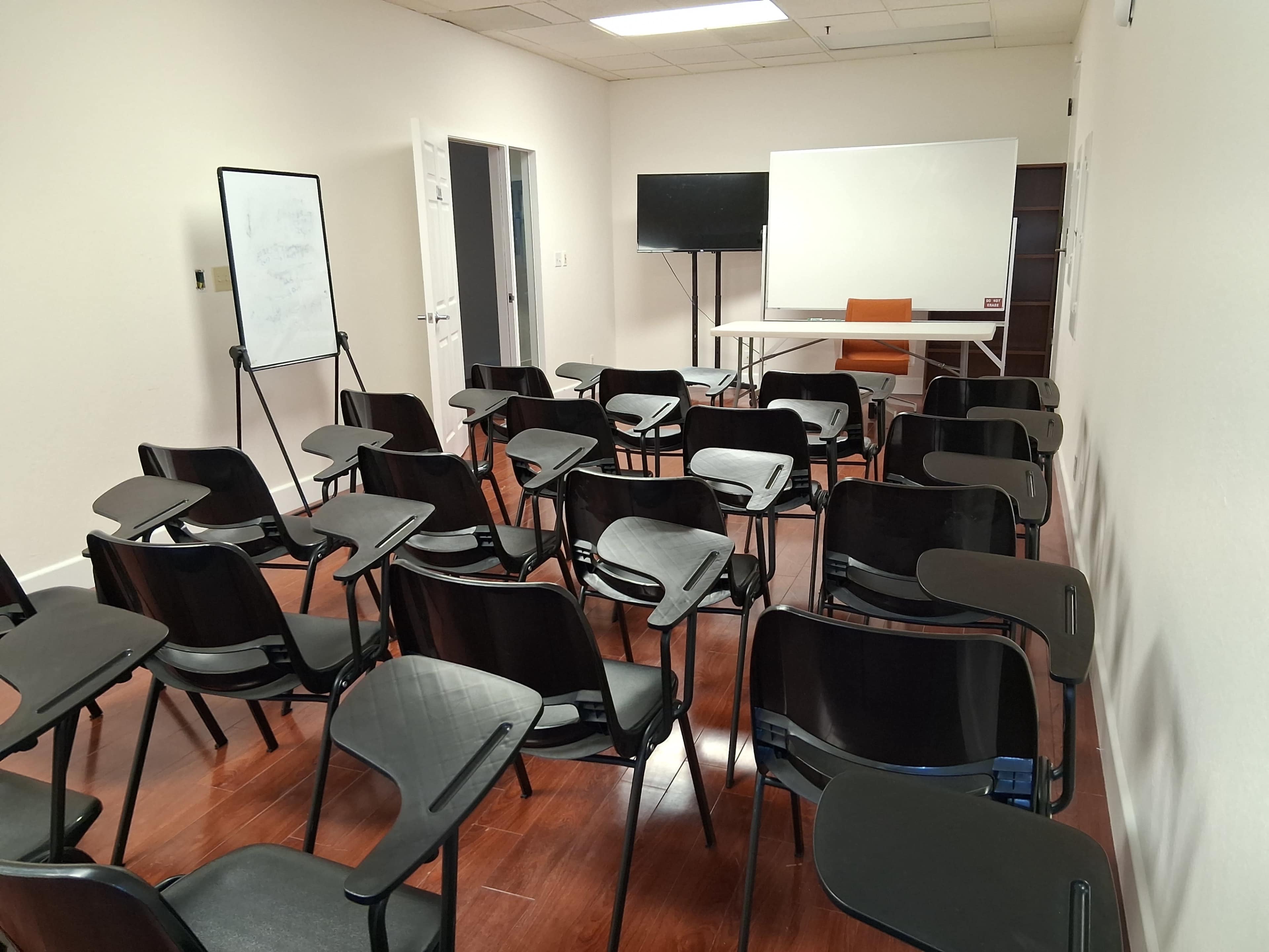 The image shows a classroom or meeting room with rows of black chairs facing a table and a presentation screen.