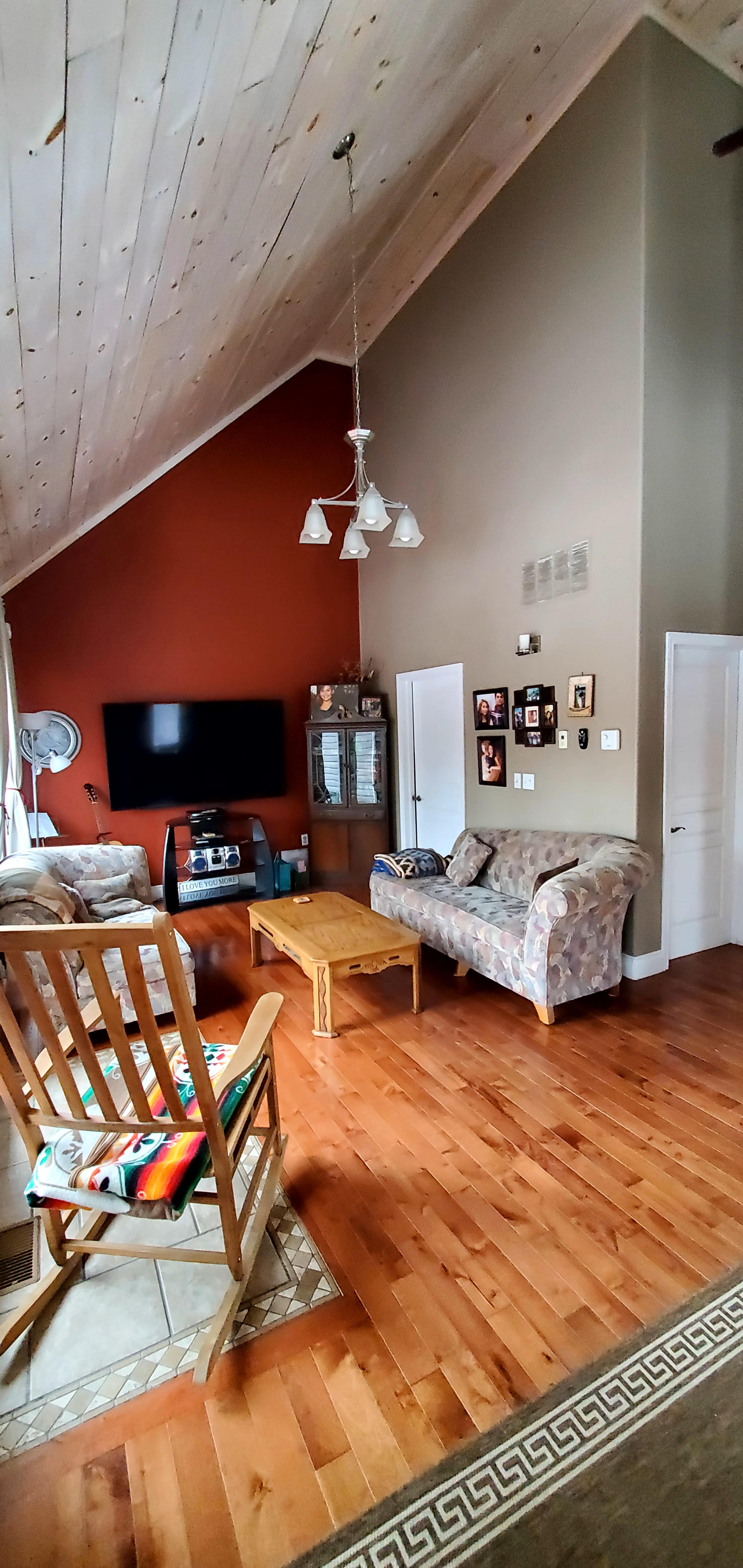The living room features a high ceiling, wooden floor, a large TV mounted on the wall, a couch, a coffee table, and a rocking chair.
