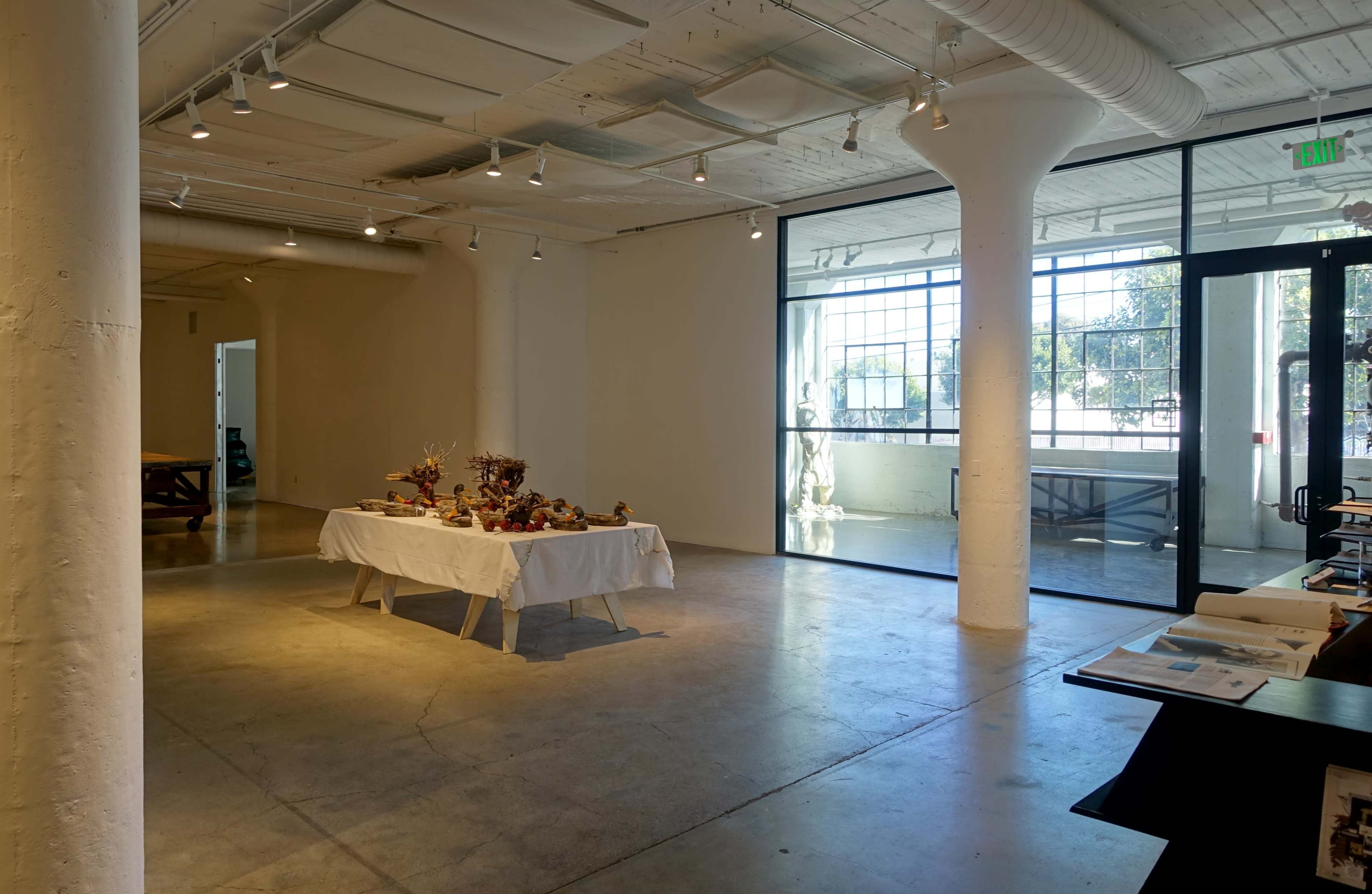 The image shows a spacious gallery with a table displaying items covered by a white cloth, alongside large windows that let in natural light.