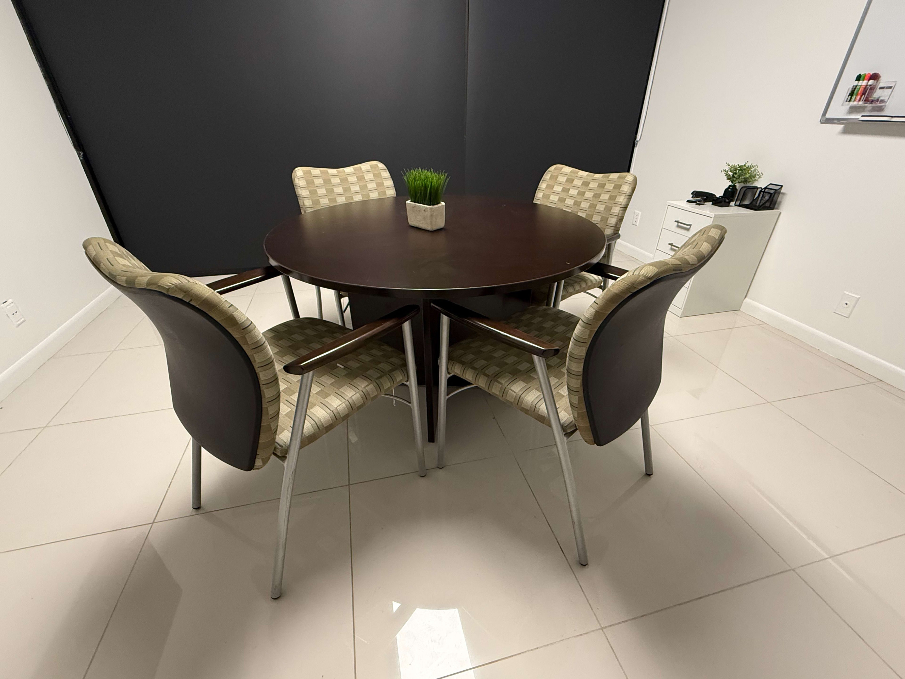 A round wooden table with four patterned chairs is set in a minimally decorated room with black walls.