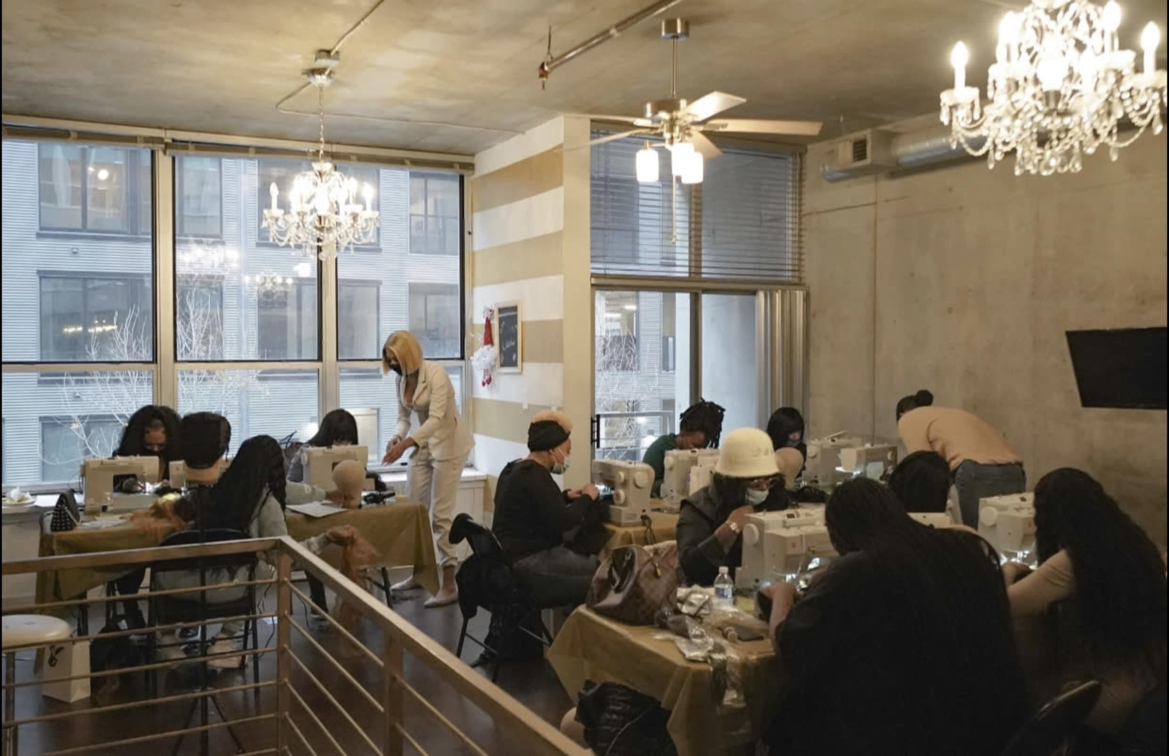 A group of people is engaged in a sewing workshop, using sewing machines at tables in a well-lit room with large windows.