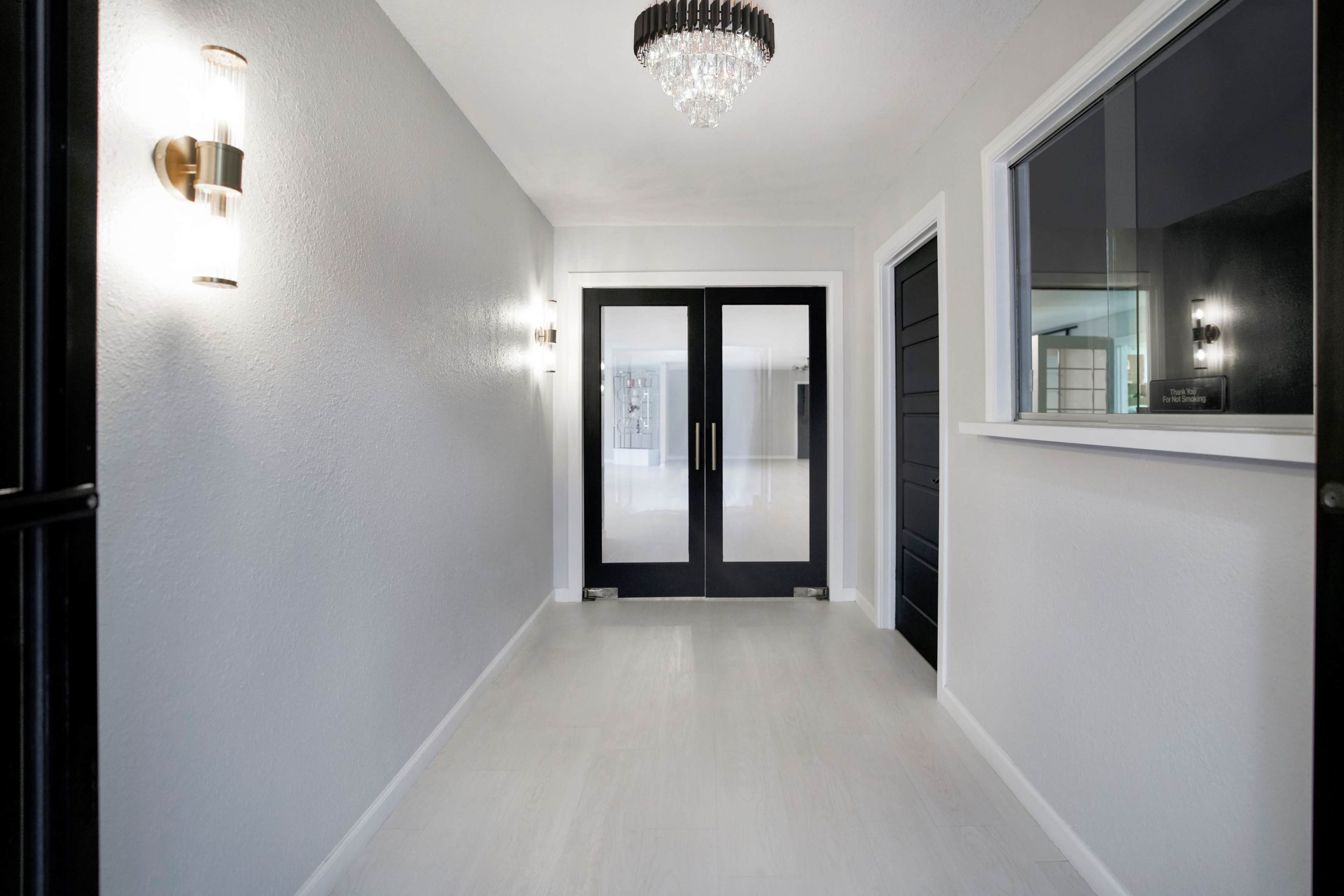 A clean, white hallway features double black doors at the end, with a chandelier overhead and a large window to the side.