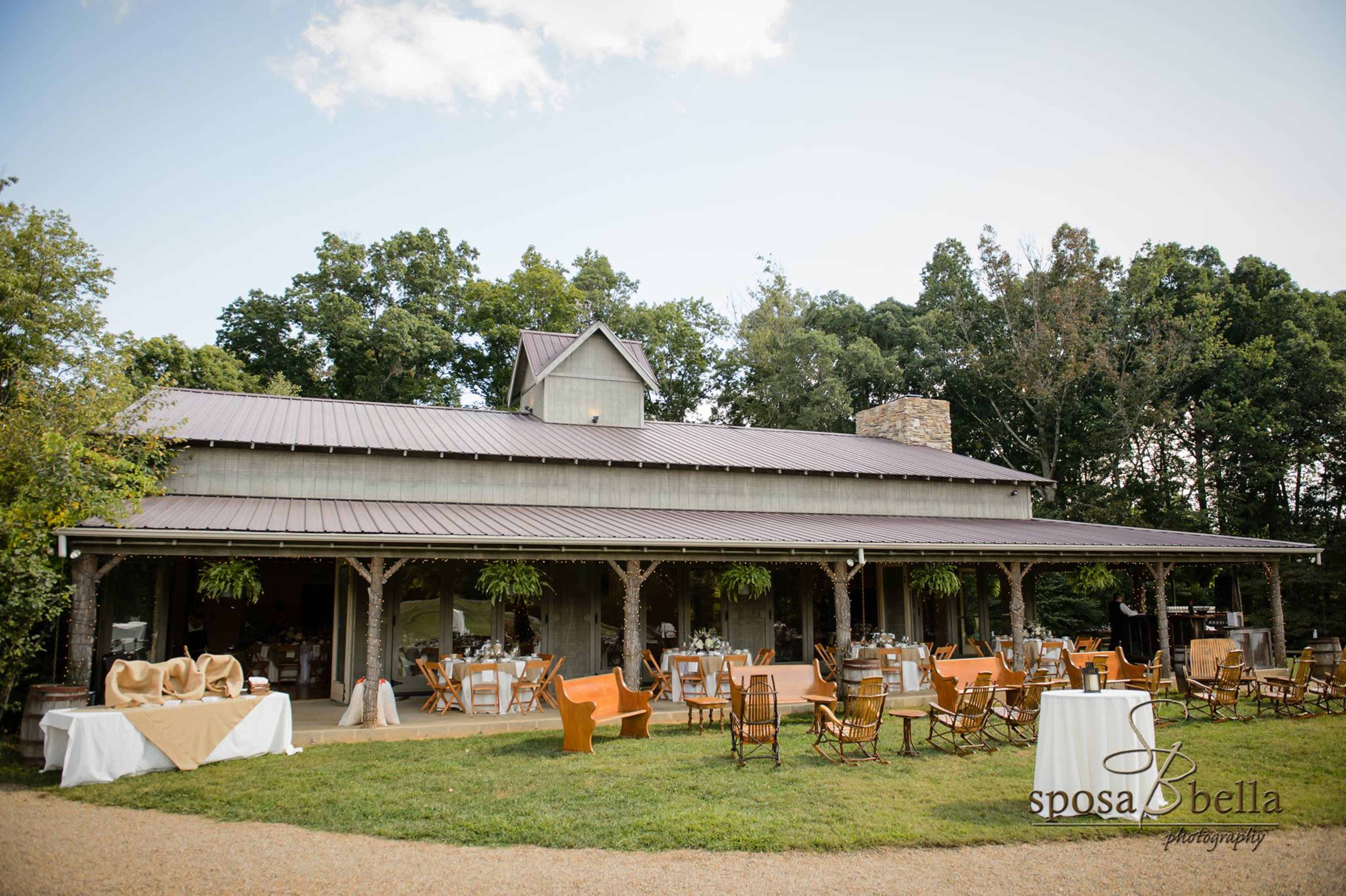 A rustic venue with a spacious porch, set up for an outdoor event, featuring tables and chairs arranged on a gravel path.