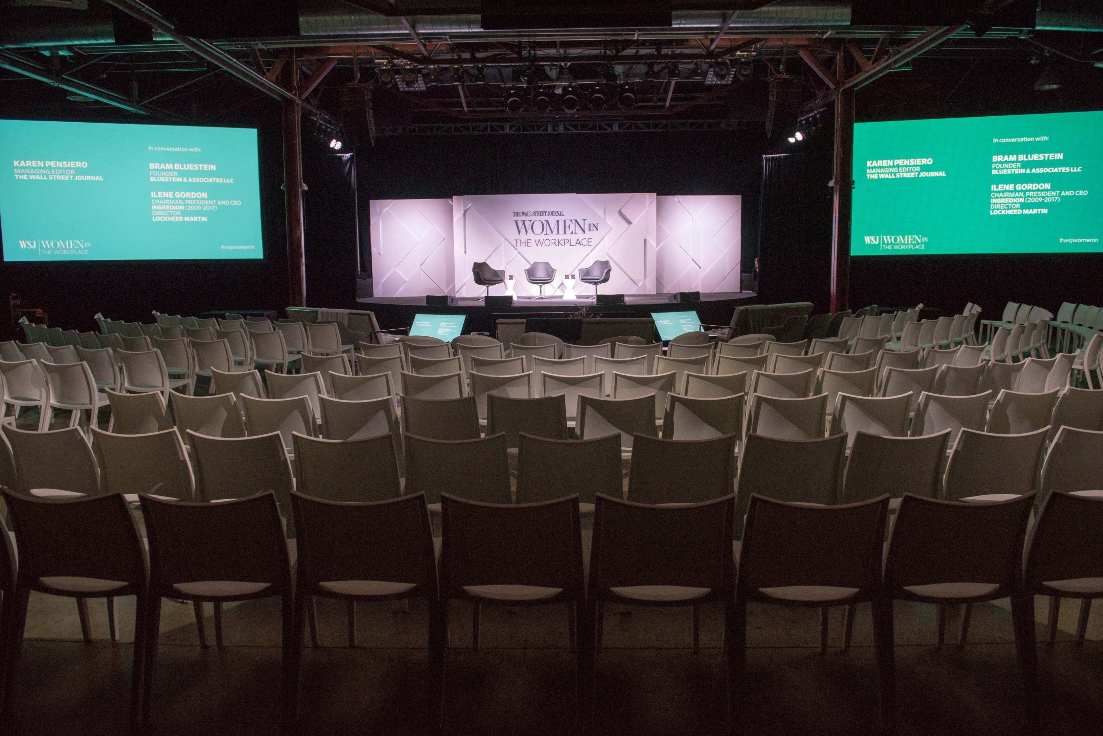 An auditorium set up for a "Women in the Workplace" event, featuring empty rows of chairs facing a stage with large screens on either side.