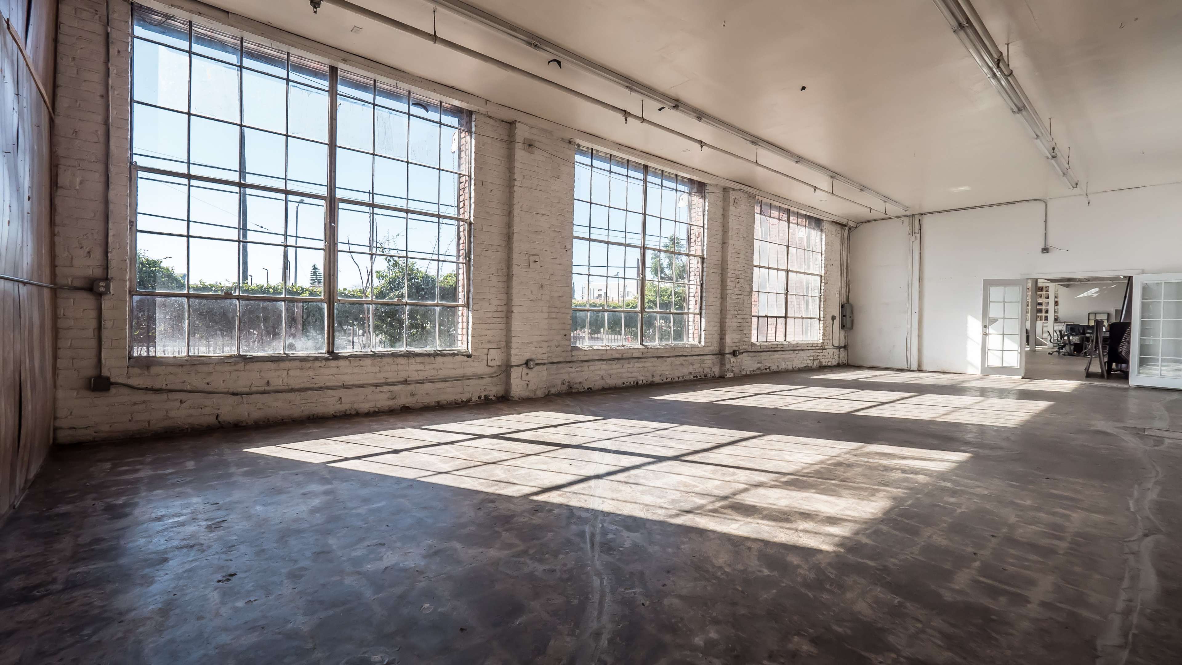 The image shows a spacious, empty room with large windows allowing natural light to flood in, casting shadows on the smooth concrete floor.
