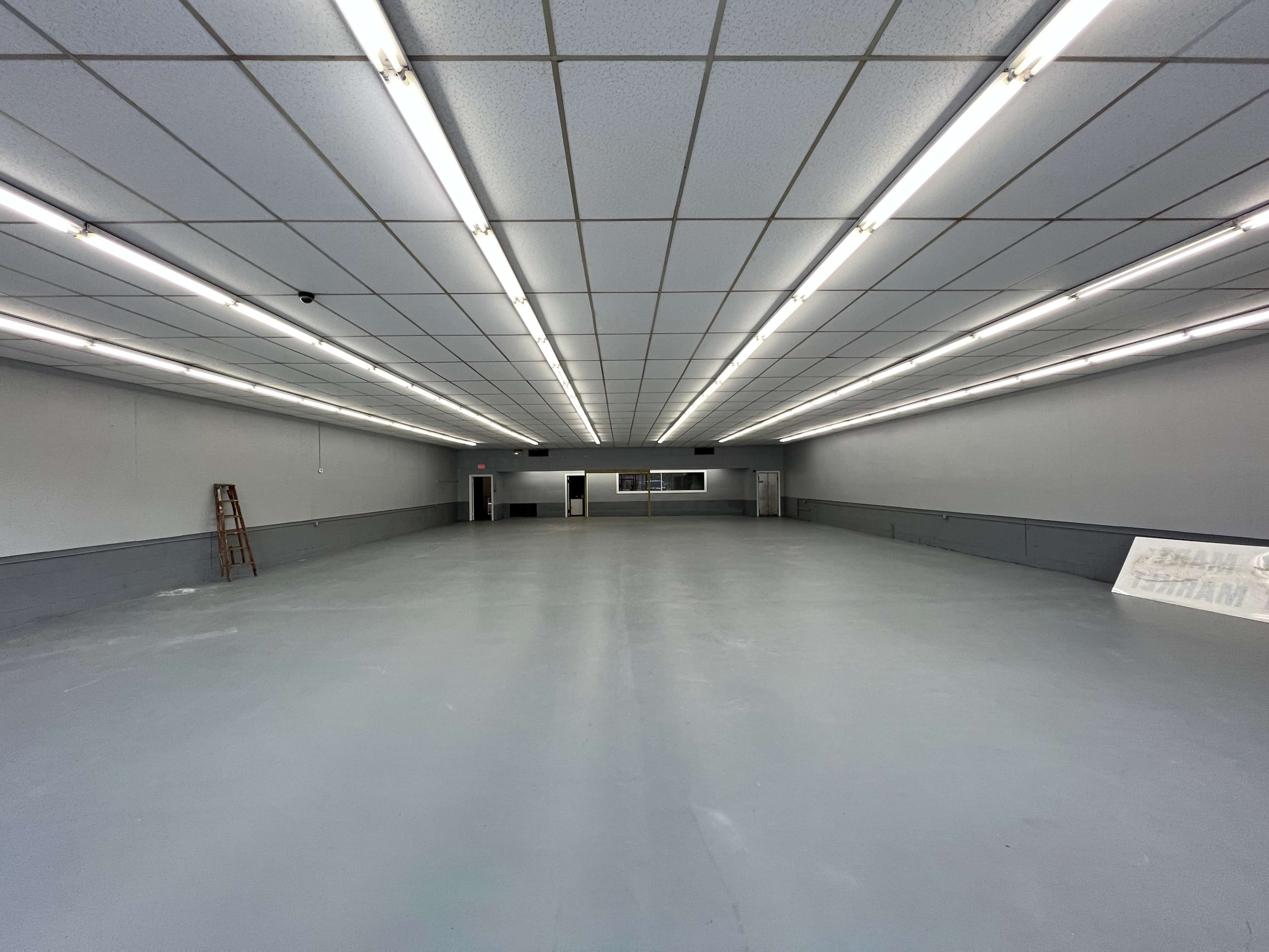 A large, empty interior space with gray flooring, bright overhead lights, and plain walls.