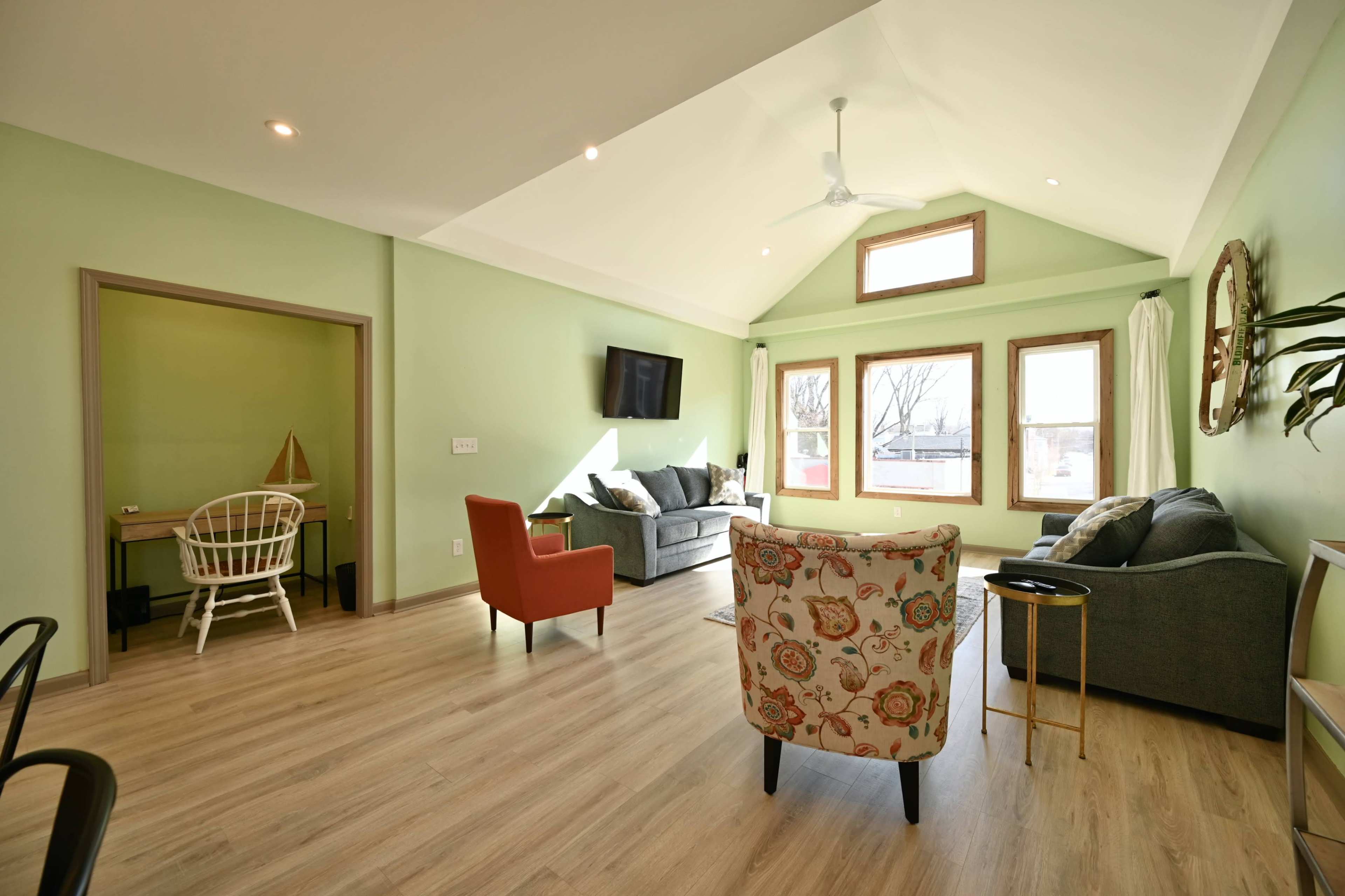 A modern living room with green walls, a high ceiling, and a mix of seating options, including a colorful chair and a gray sofa.