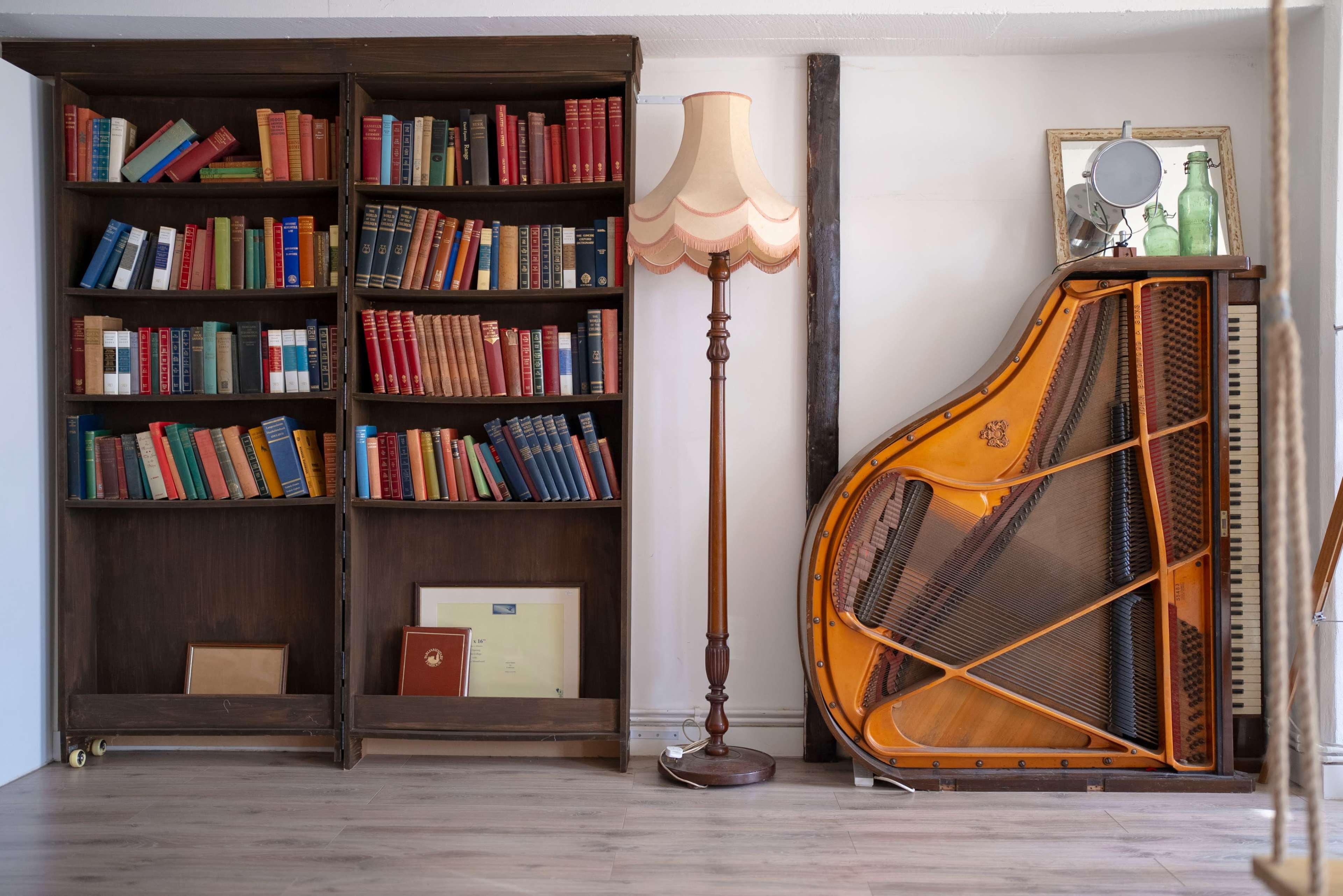 A wooden bookshelf filled with colorful books stands next to a vintage lamp and a grand piano.