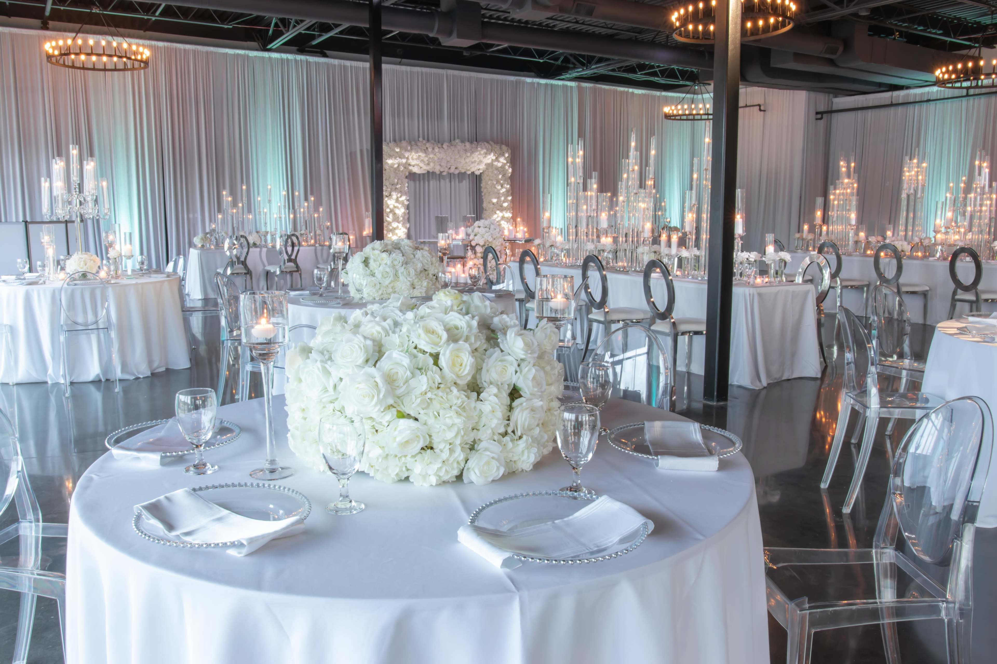 An elegantly arranged banquet hall featuring round tables with white tablecloths, clear ghost chairs, and a large floral centerpiece of white roses, illuminated by soft candlelight and decorative lighting in the background.