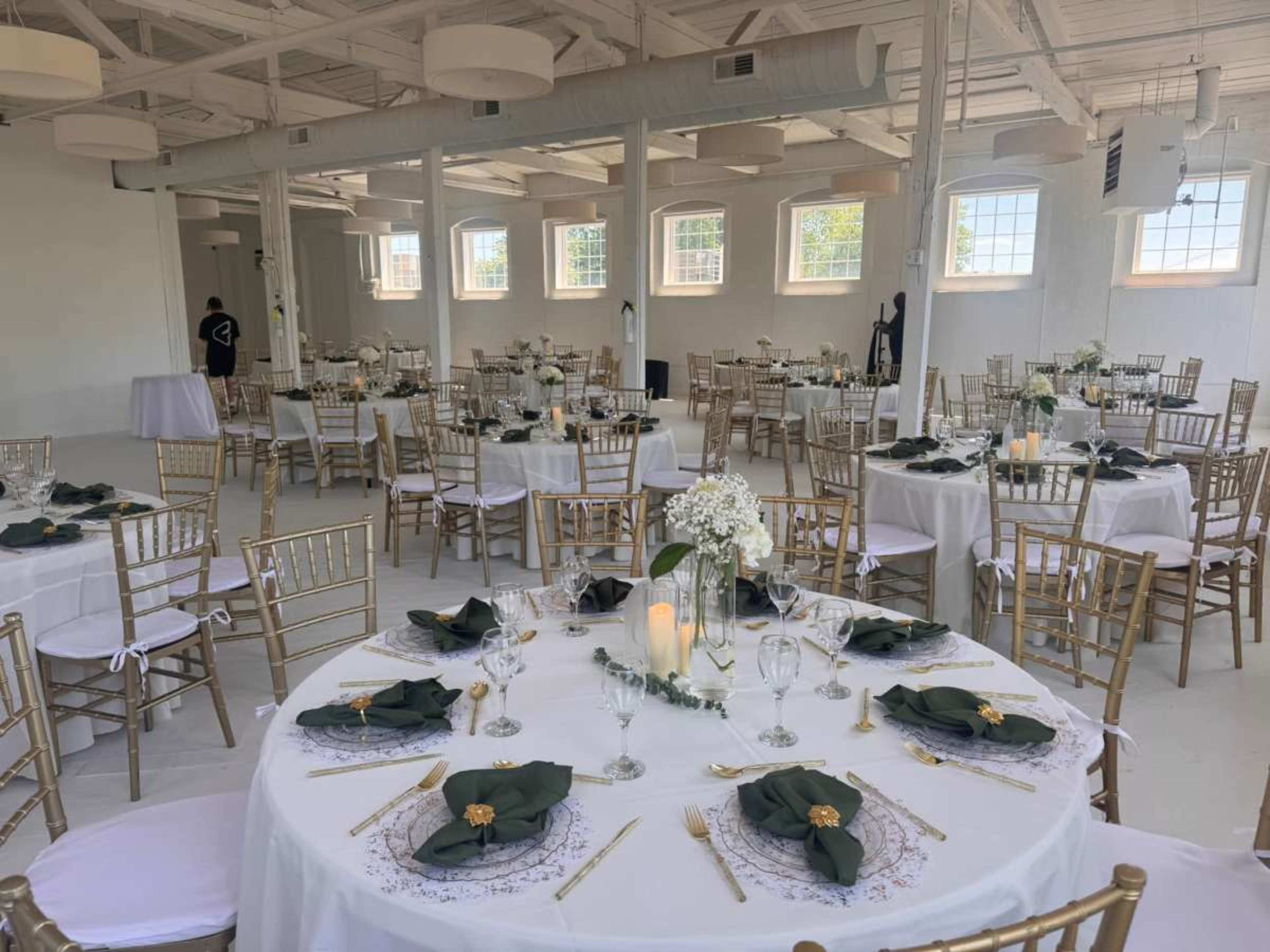 The image shows an elegantly arranged dining hall set for a wedding, featuring multiple round tables with white tablecloths, gold chairs, and floral centerpieces.