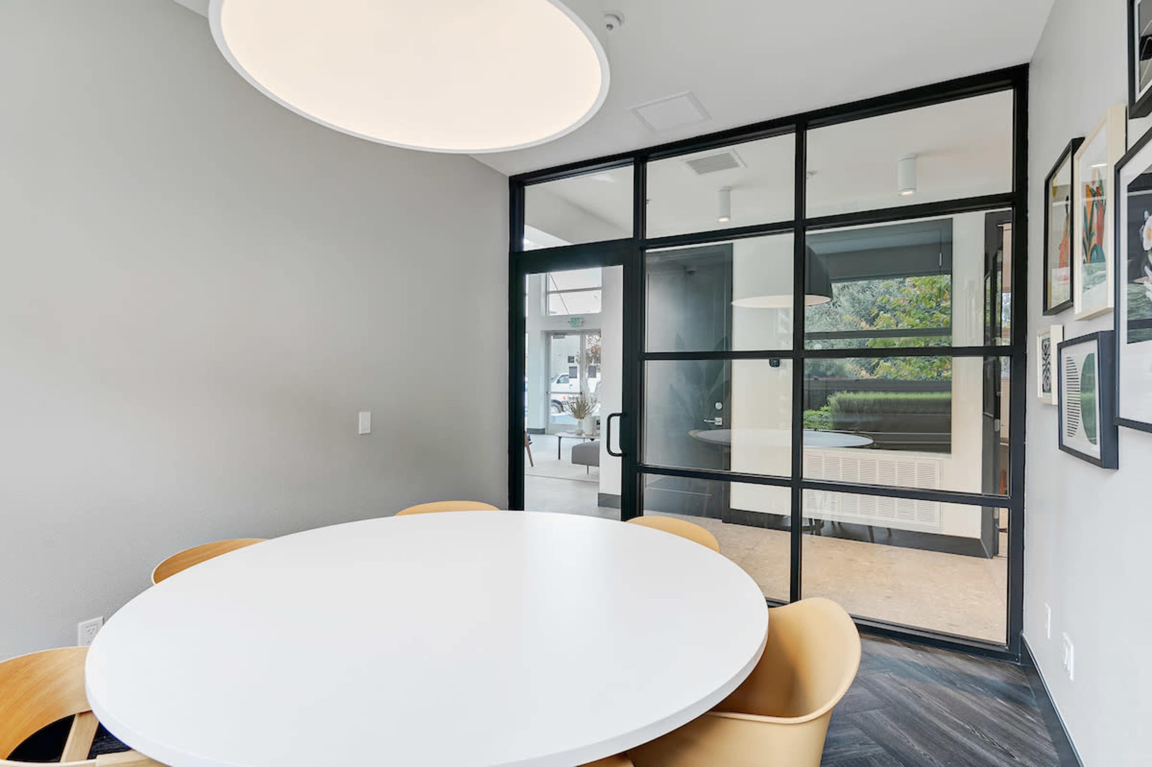 A dining area features a round white table surrounded by orange chairs, with large glass doors leading to an outdoor view.