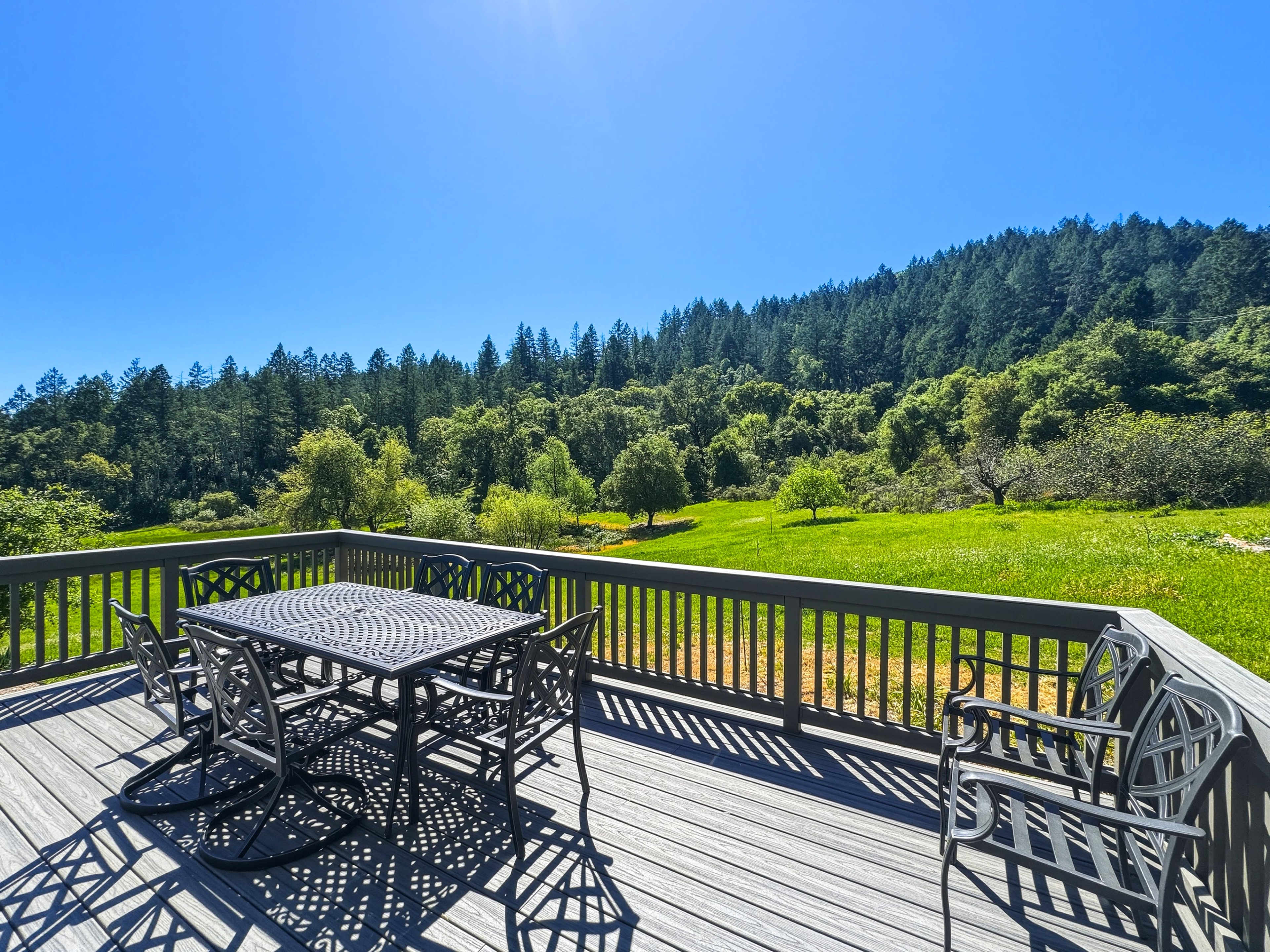 A deck with metal outdoor furniture overlooks a lush green landscape filled with trees and rolling hills under a clear blue sky.