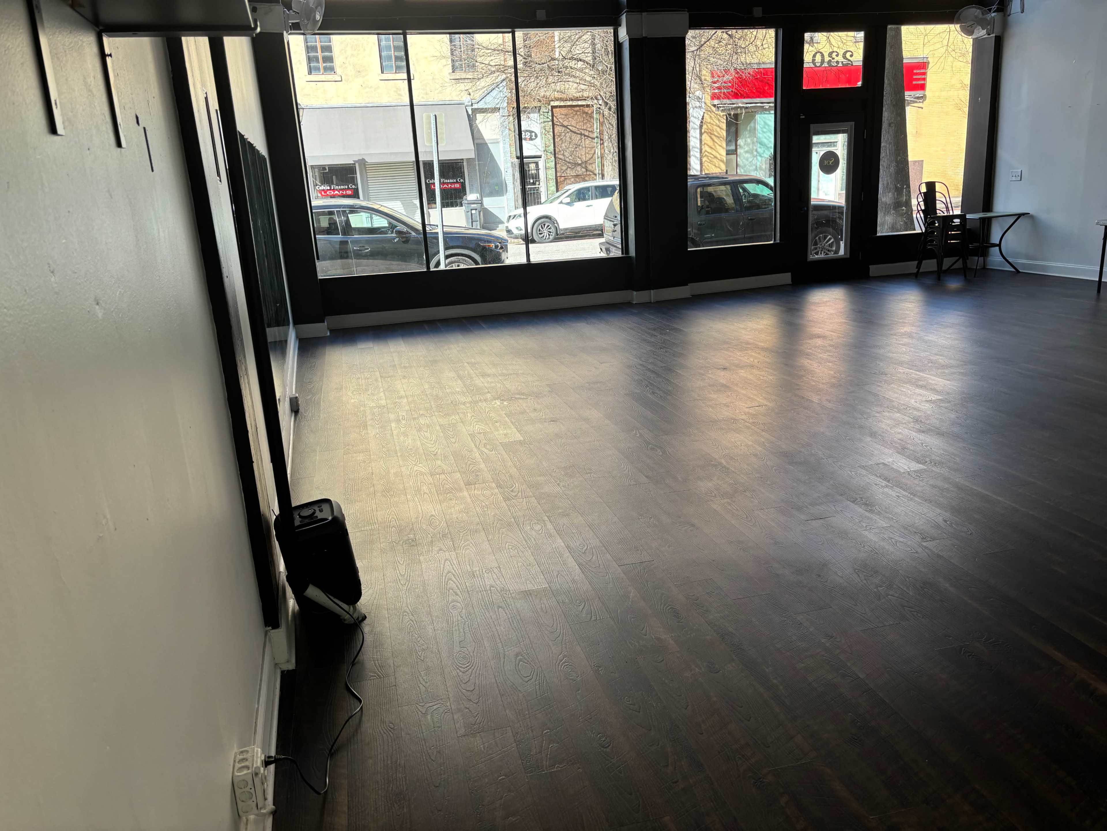 A spacious, empty interior with dark wooden floors and large windows facing an urban street.