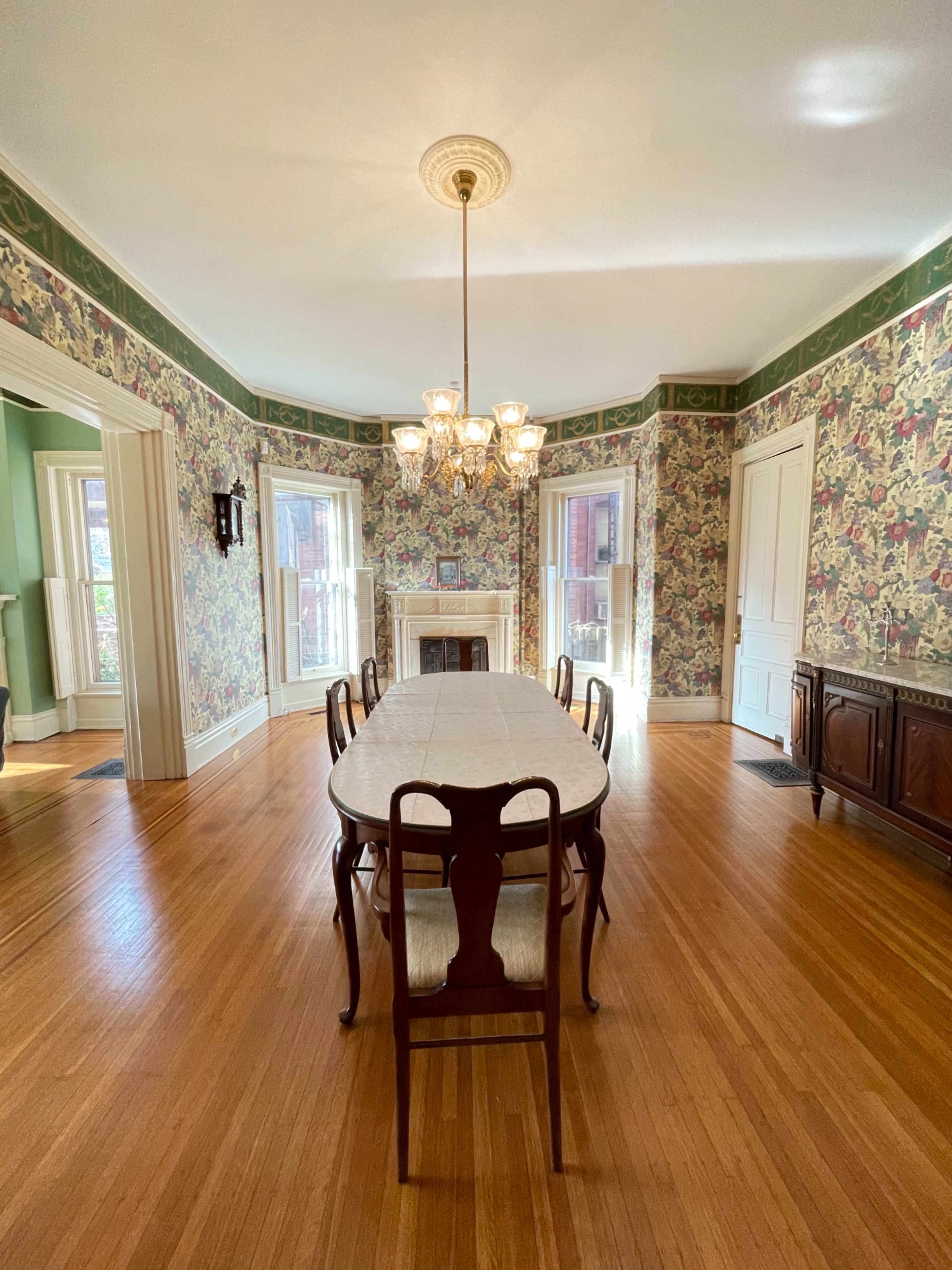 A dining room with a long table surrounded by chairs, adorned with floral wallpaper and a chandelier overhead.