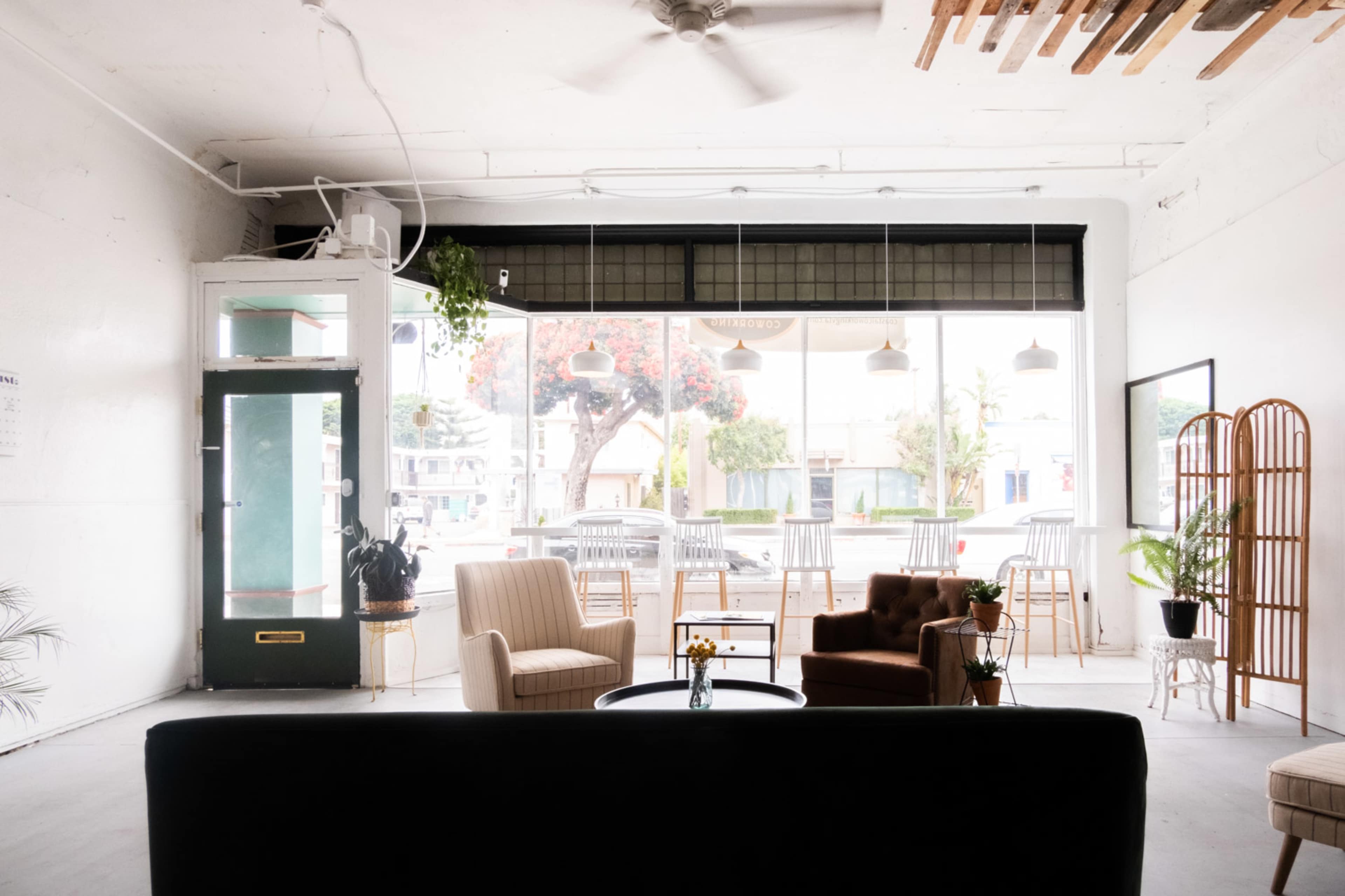 The image shows a spacious, open room with large windows, featuring a seating area with two armchairs, a coffee table, and a view of outdoor seating.