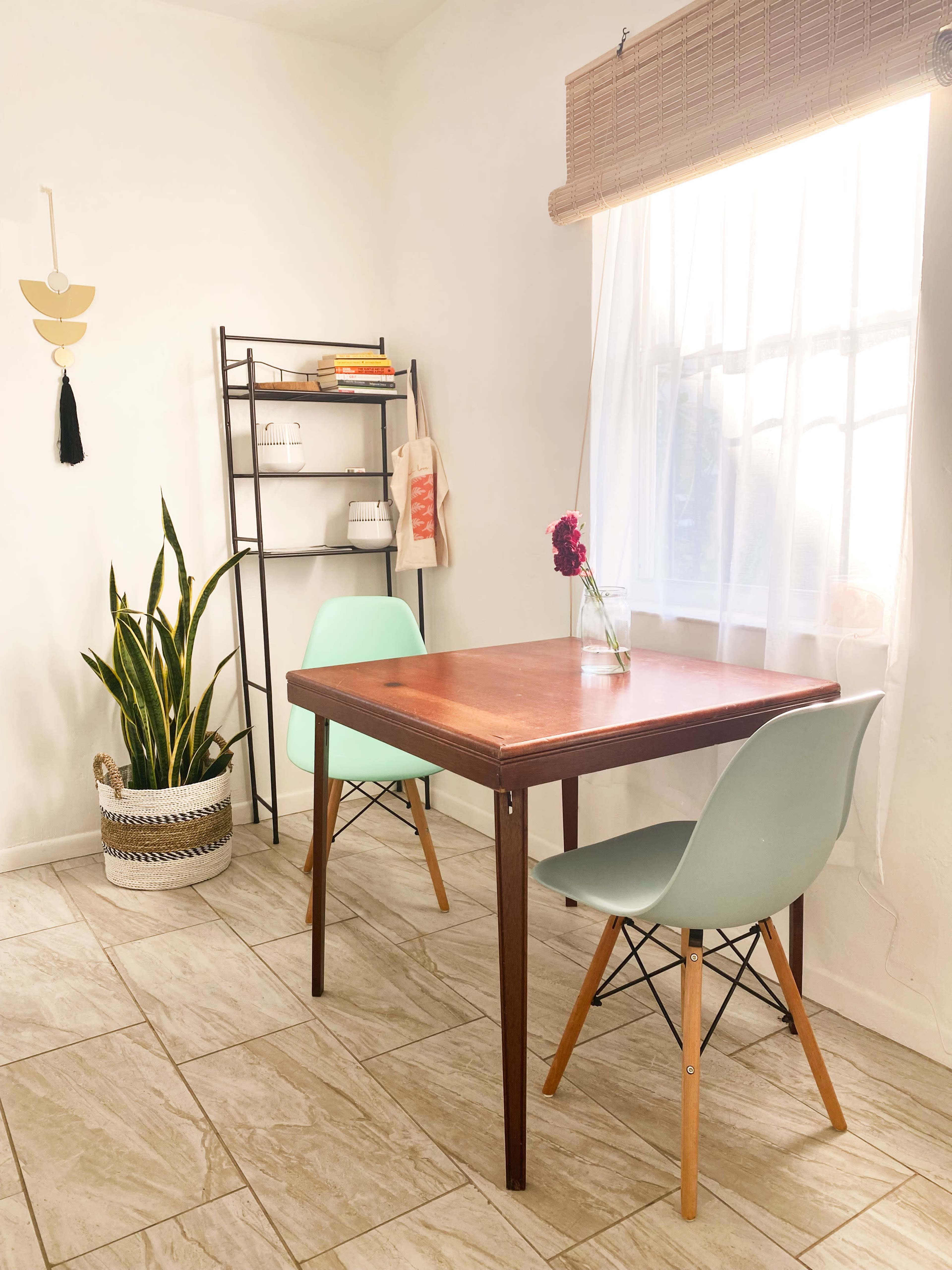 A wooden table is set between two light green chairs in a room featuring a large window, a plant in a woven pot, and a bookshelf in the background.