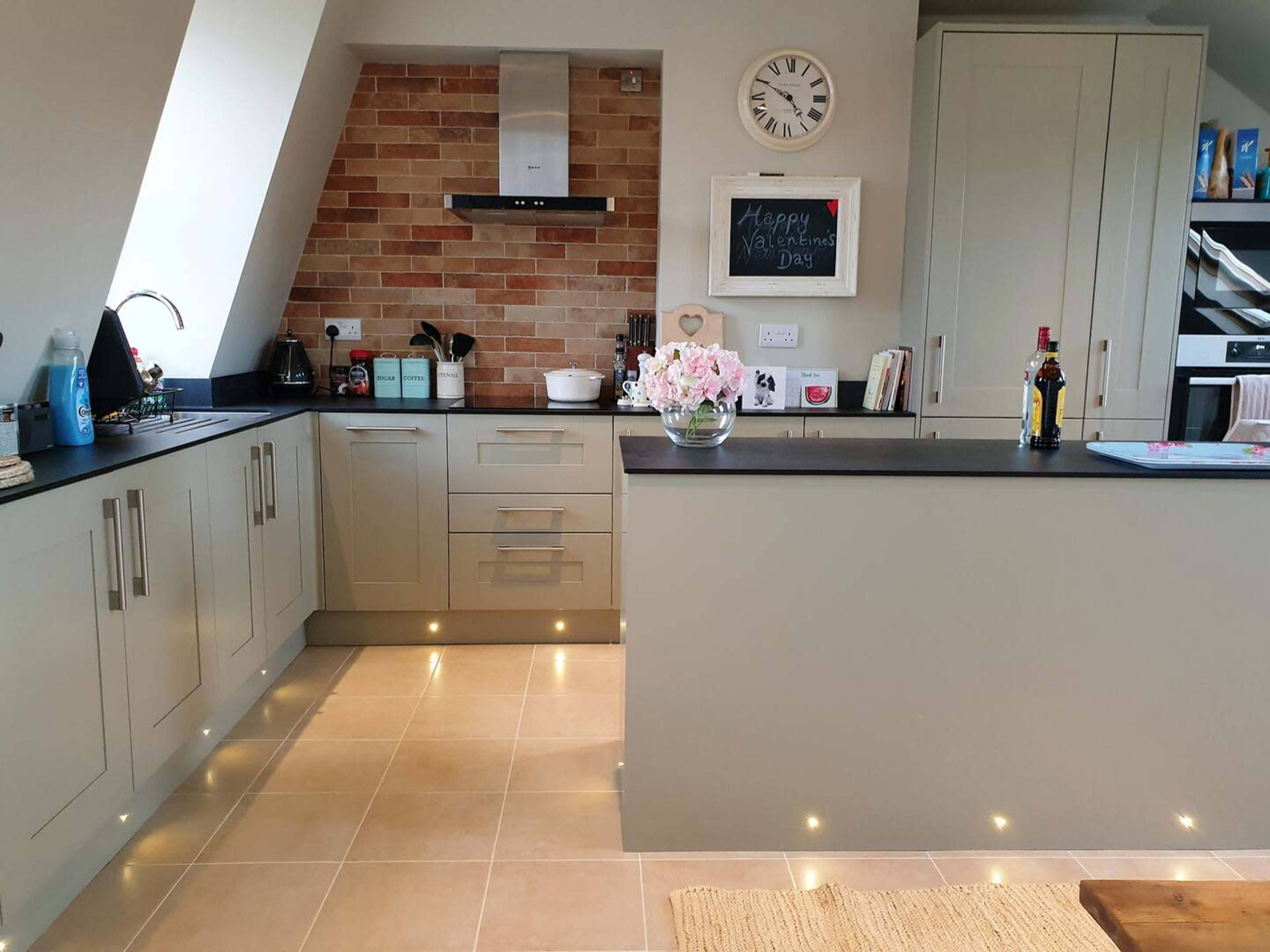 The image shows a modern kitchen with light gray cabinetry, a brick backsplash, and a central island, featuring a vase of flowers and various kitchen appliances.
