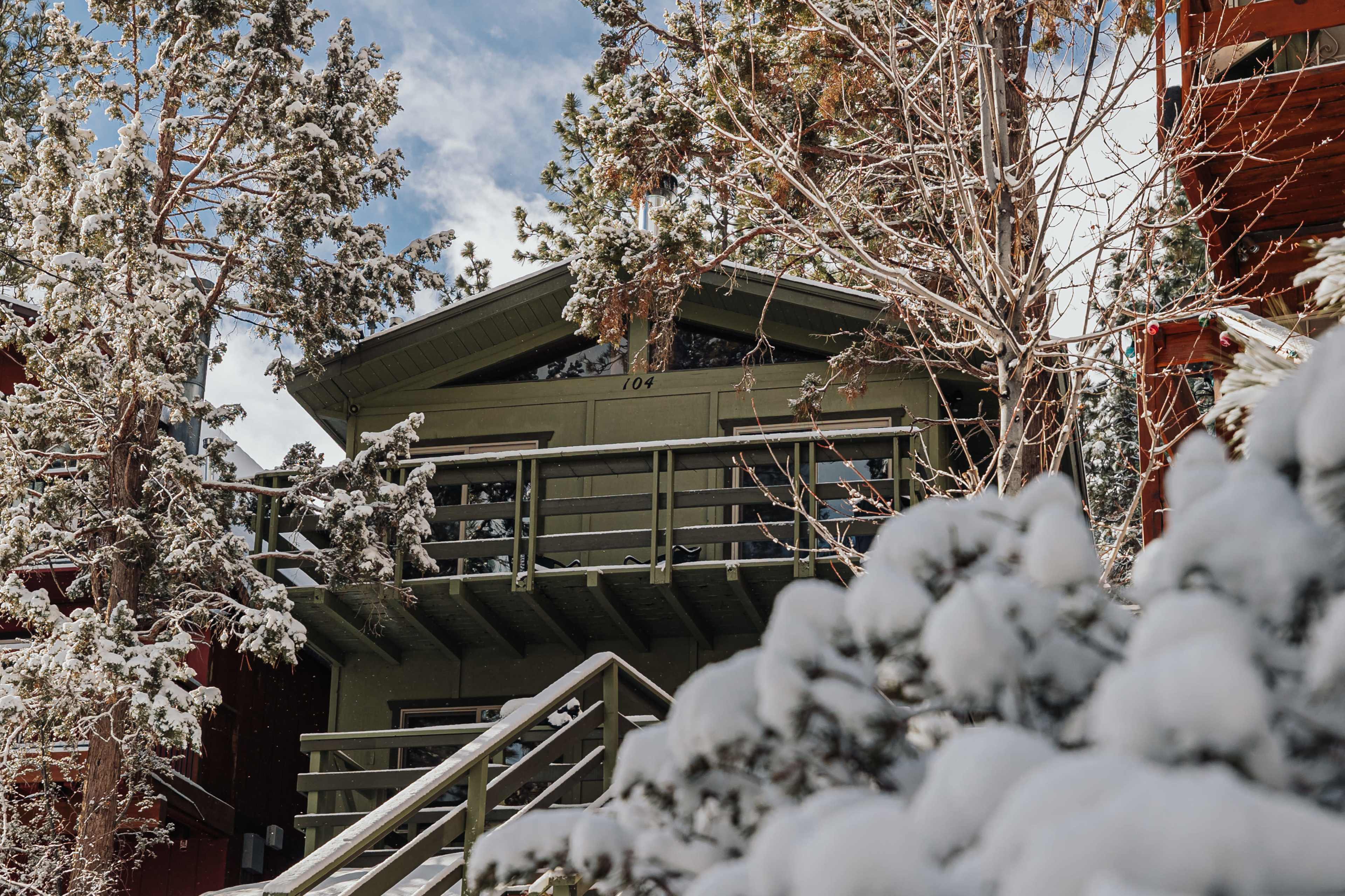 A snowy scene features a green cabin with a balcony, nestled among trees in a winter landscape.