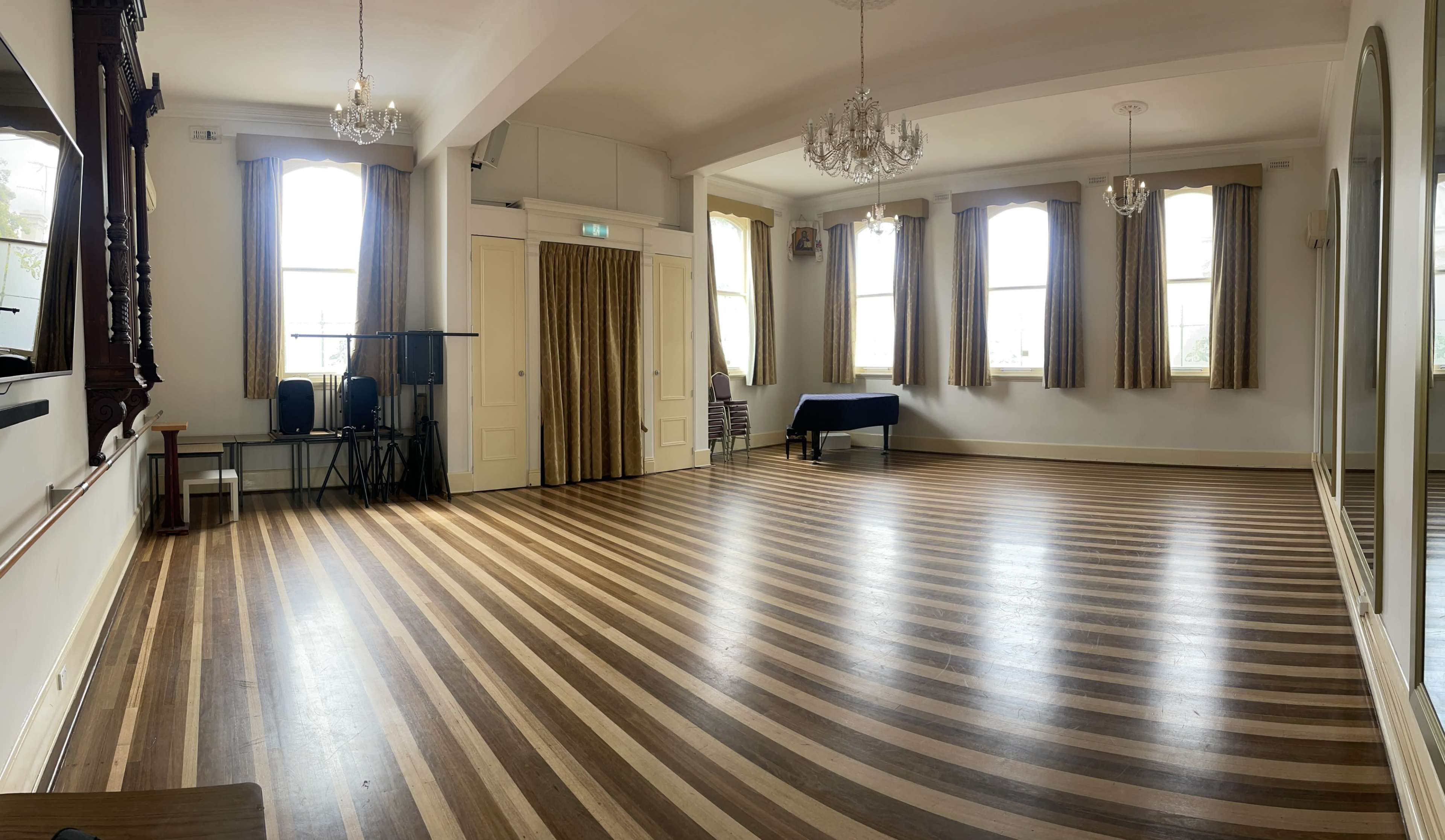 A spacious, empty room with polished wooden floors and large windows, featuring cream-colored walls and two chandeliers.