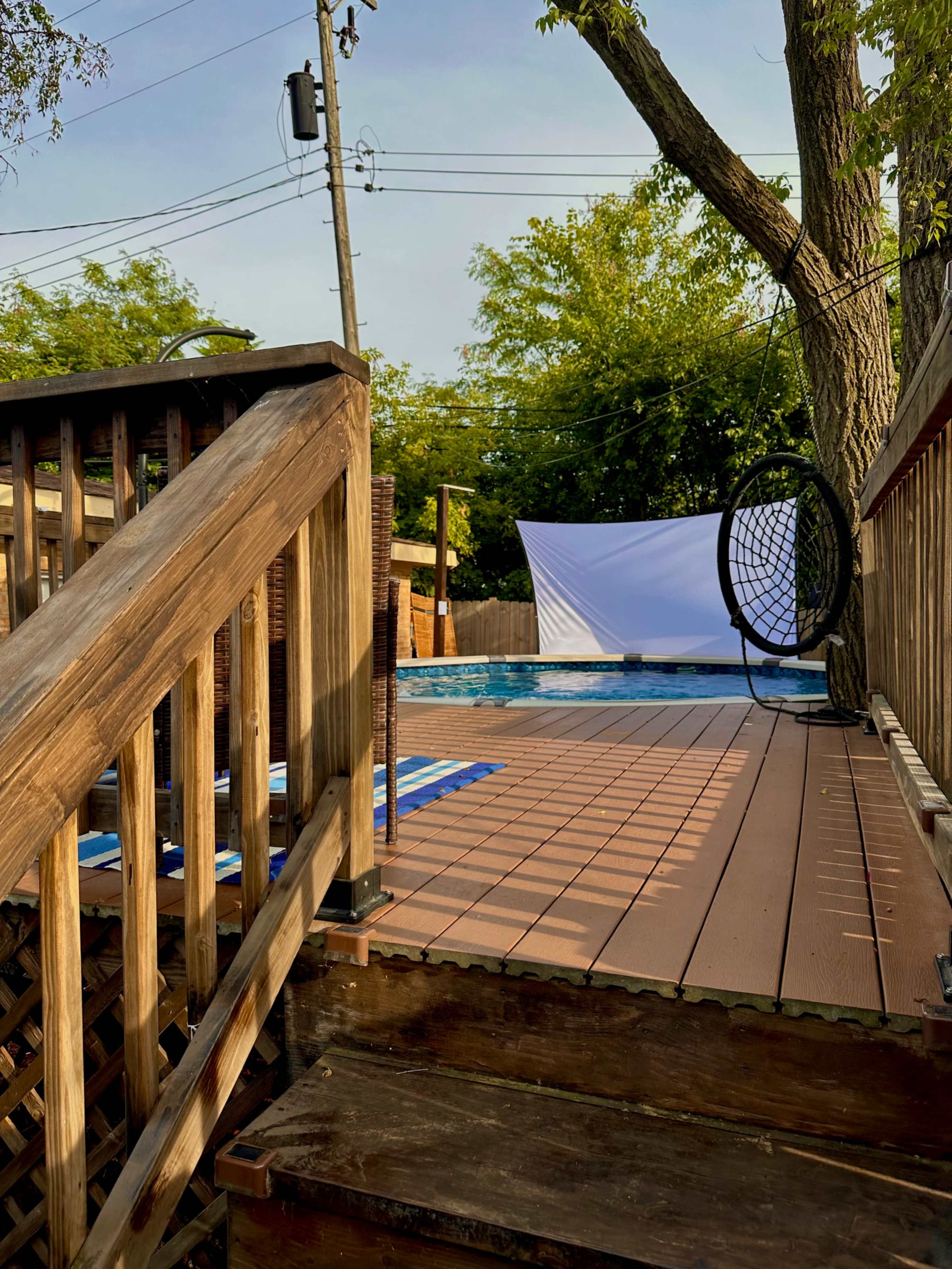 A wooden deck leads down to a pool area surrounded by trees and a white backdrop.