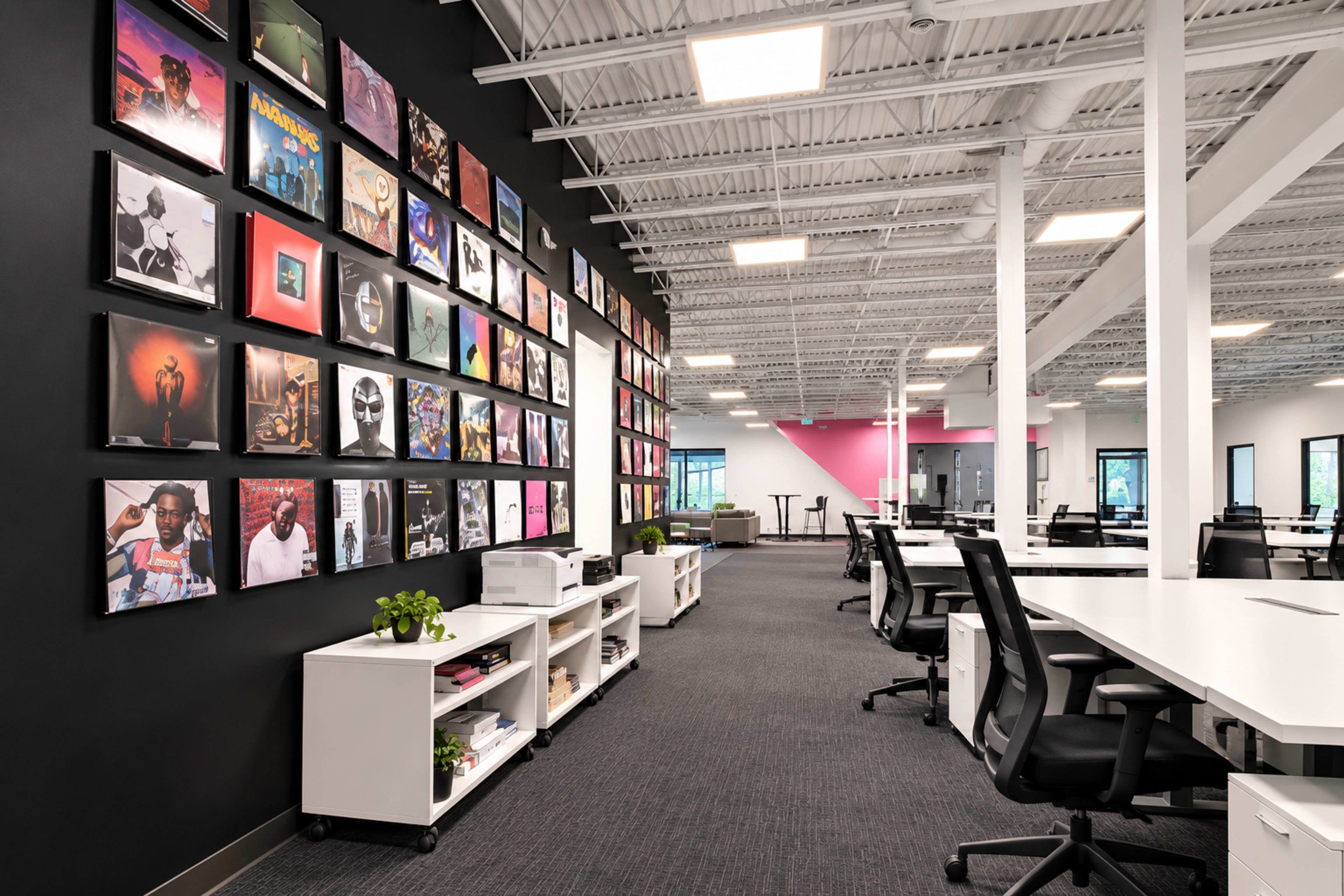 The image shows a modern office space with white desks, black and pink accent walls, and a gallery of framed artwork on one wall.