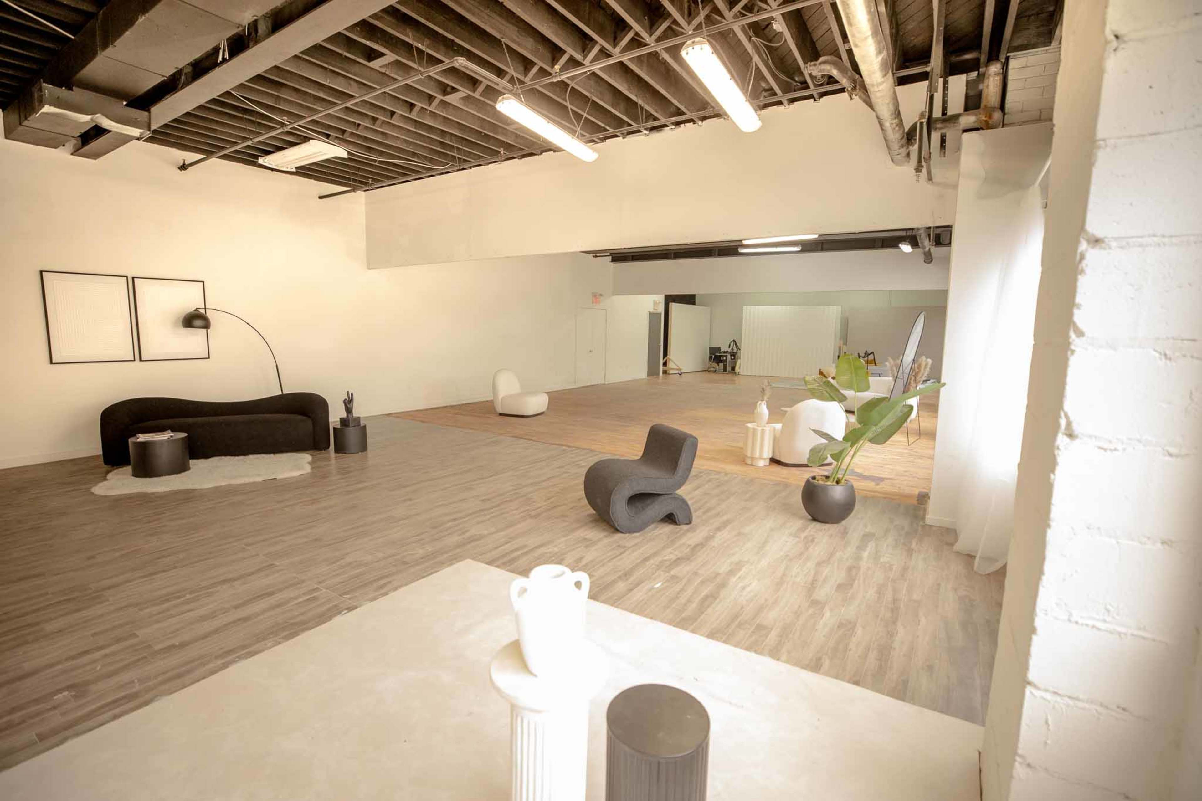 The image shows a spacious, minimalistic interior with a wooden floor, featuring modern furniture pieces like a black sofa, unique chairs, and decorative plants.