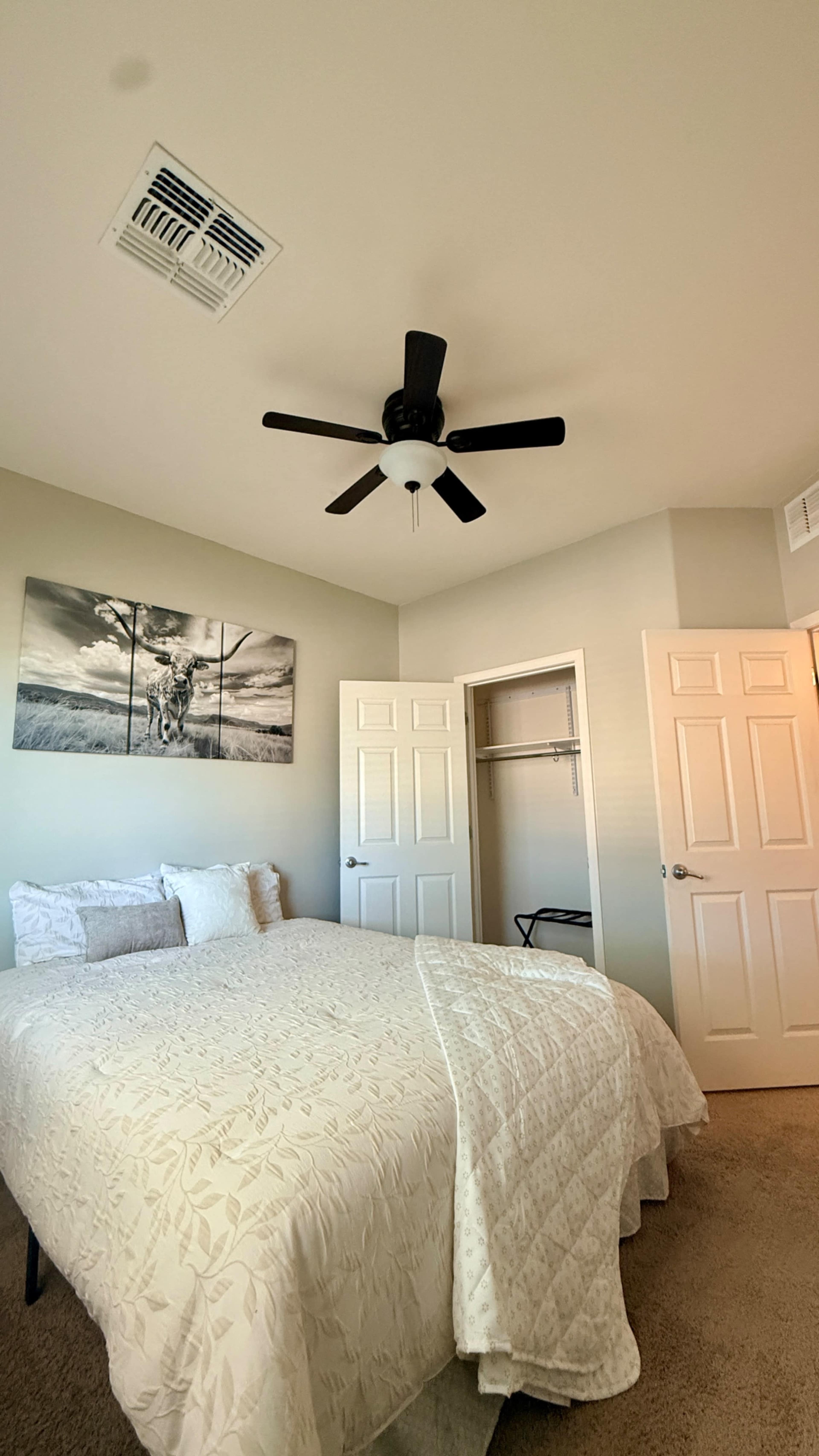 The image shows a bedroom with a bed covered in a light quilt, a ceiling fan, a closet with a hanging rod, and a wall hanging featuring a landscape.