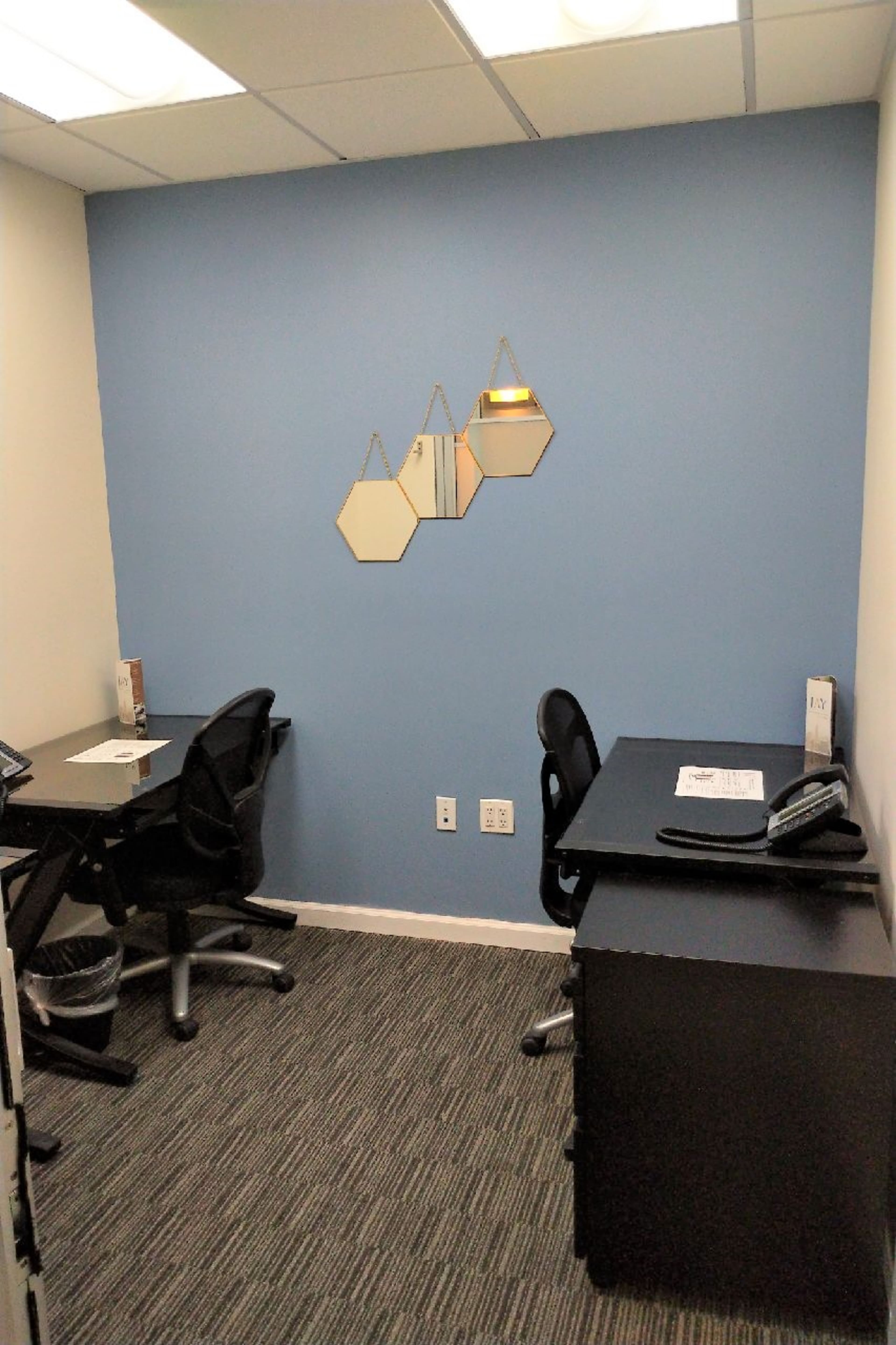 A small office space with two black desks, each equipped with a chair and a telephone, and a blue wall featuring three hexagonal mirrors.