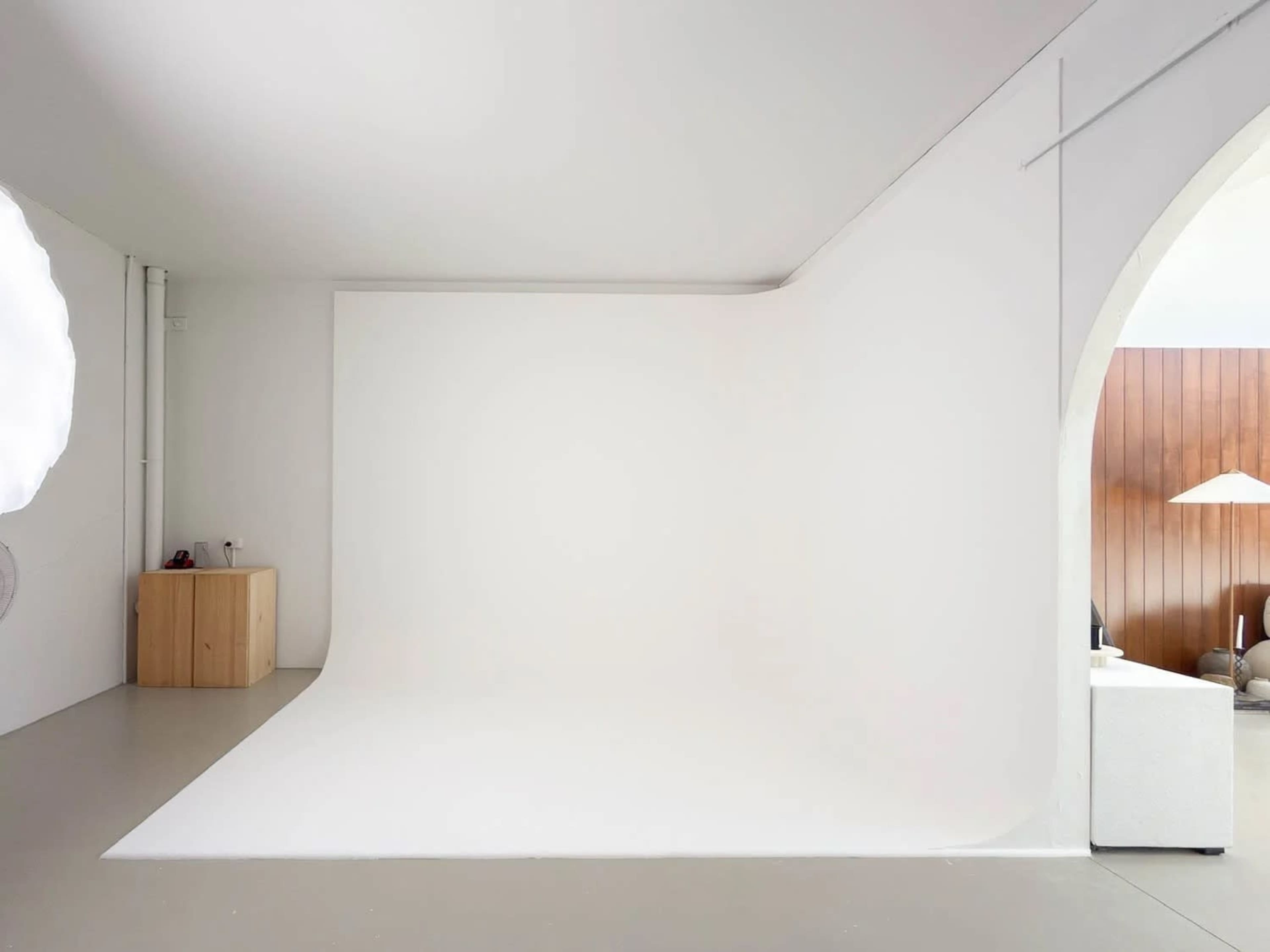 The image shows a minimalist studio space featuring a seamless white backdrop and light-colored flooring, with an archway leading to a separate area.