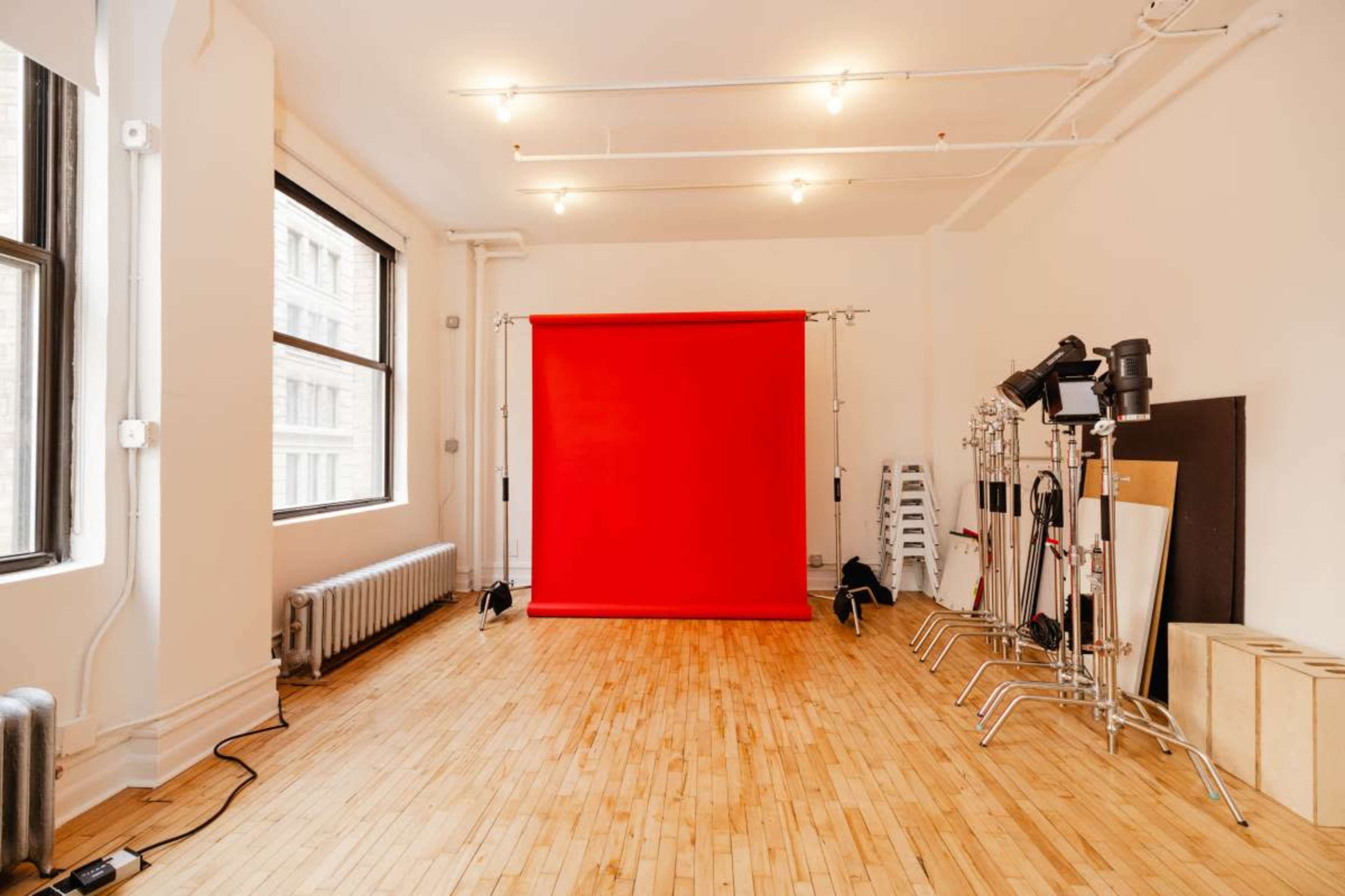 The image shows a bright studio space featuring a red backdrop, wooden flooring, and various lighting equipment arranged along the walls.