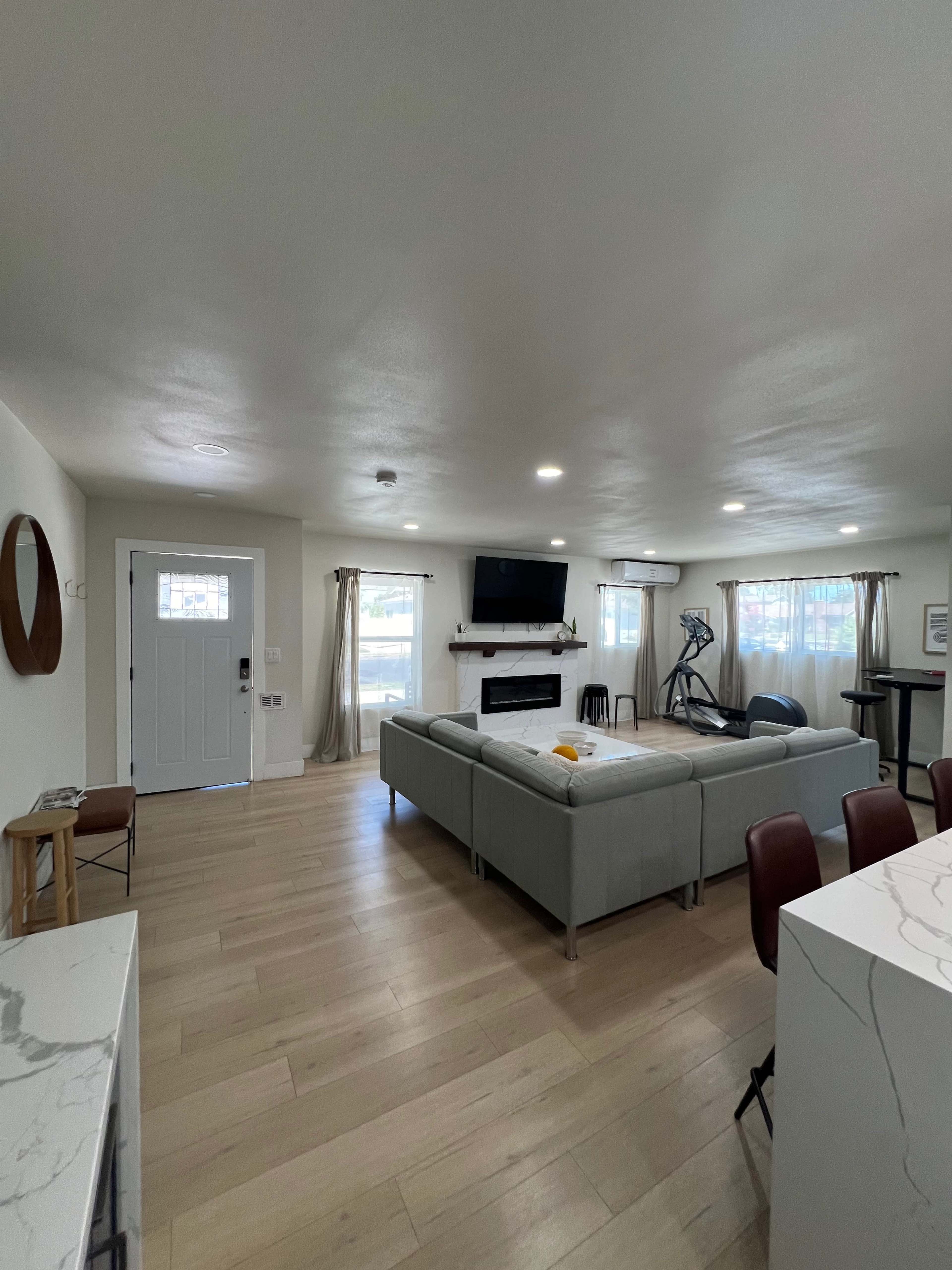 A spacious, modern living room with a light-colored sofa, a wall-mounted TV, and large windows letting in natural light.