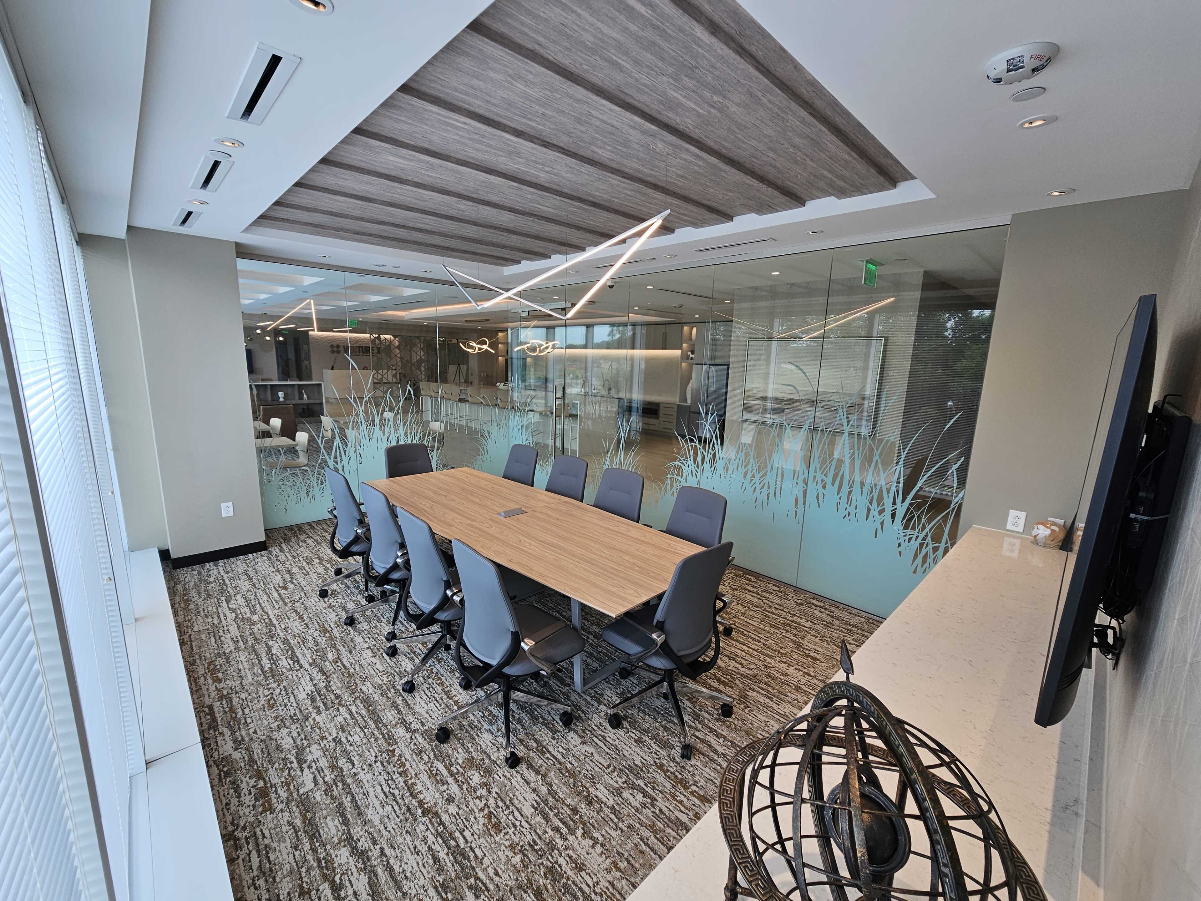 A modern conference room with a large wooden table surrounded by gray chairs, featuring glass walls and a ceiling with decorative lighting.