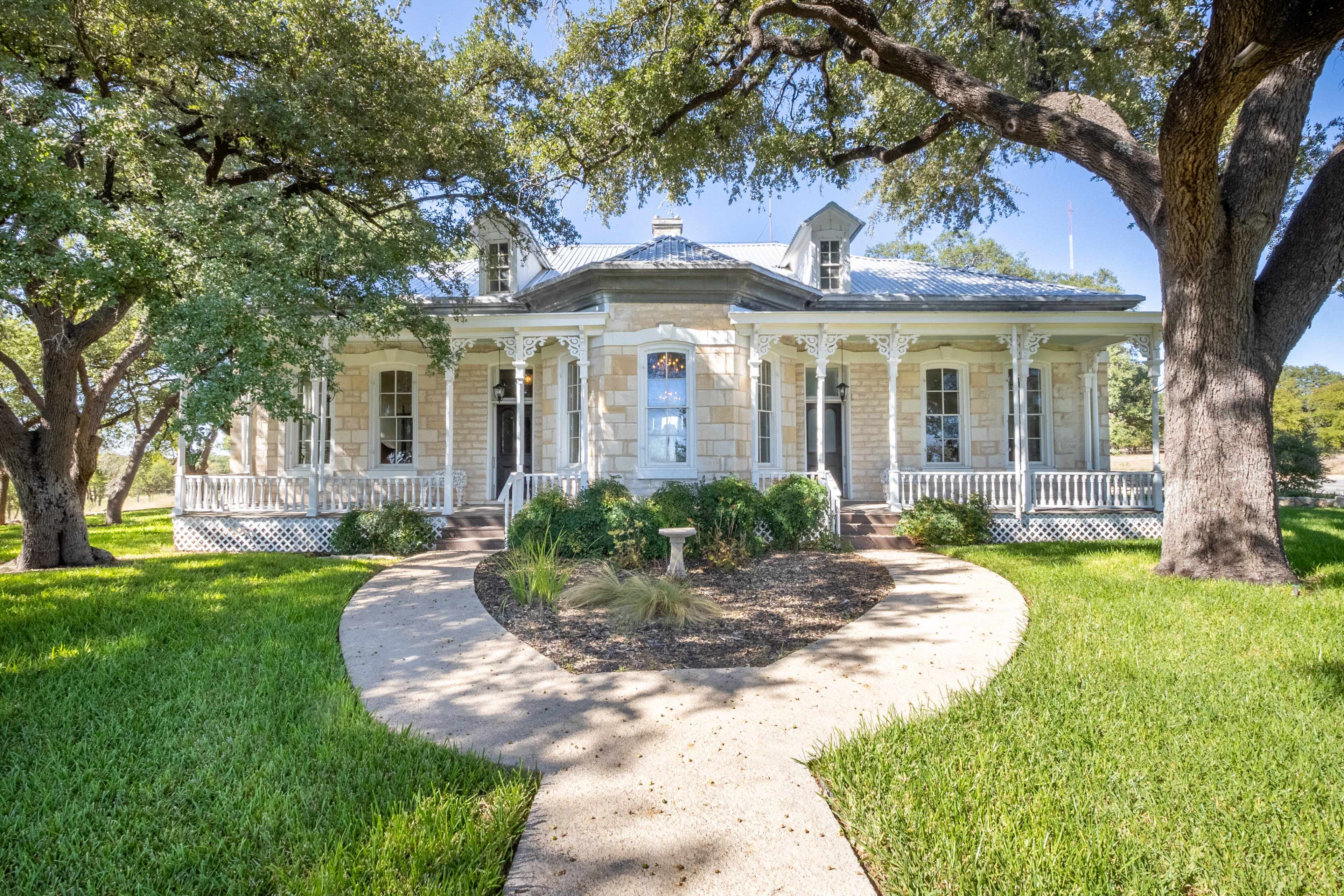 A large stone house with a front porch is surrounded by lush green grass and large trees, with a curved walkway leading up to the entrance.