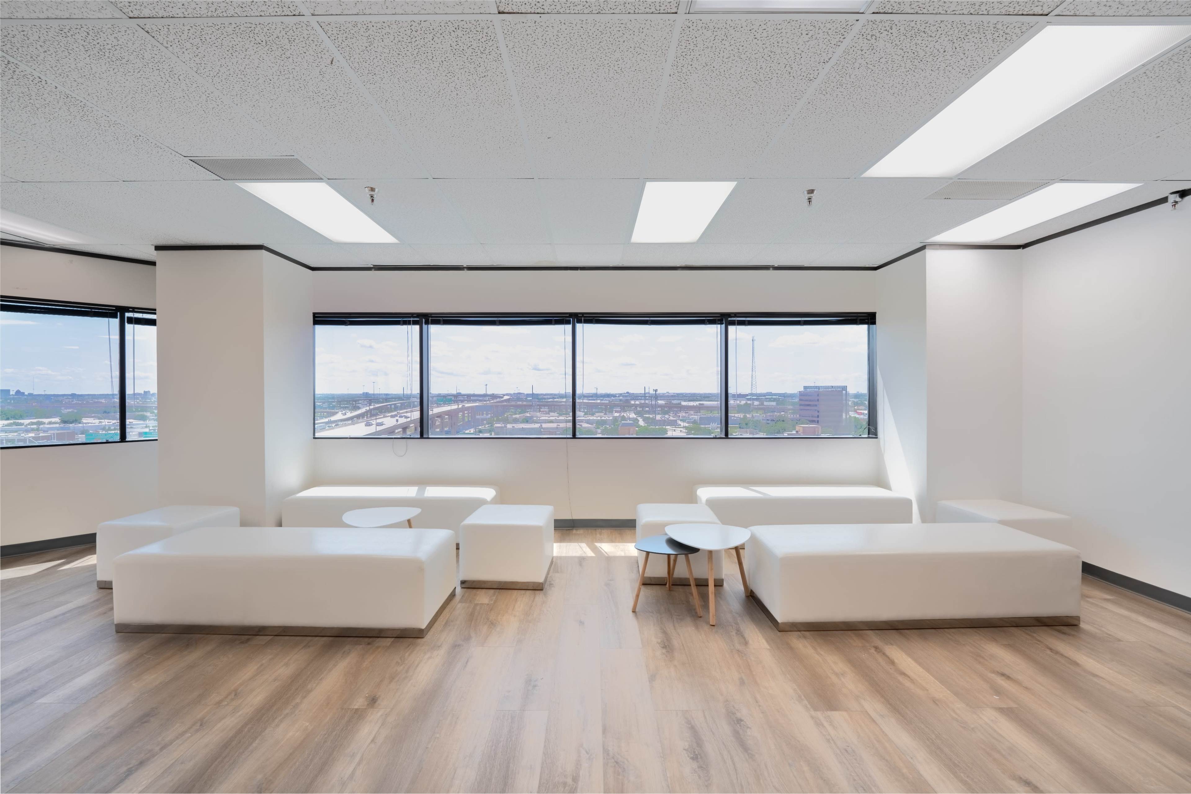 A modern office space with white seating, a small wooden table, and large windows offering a view of the city.