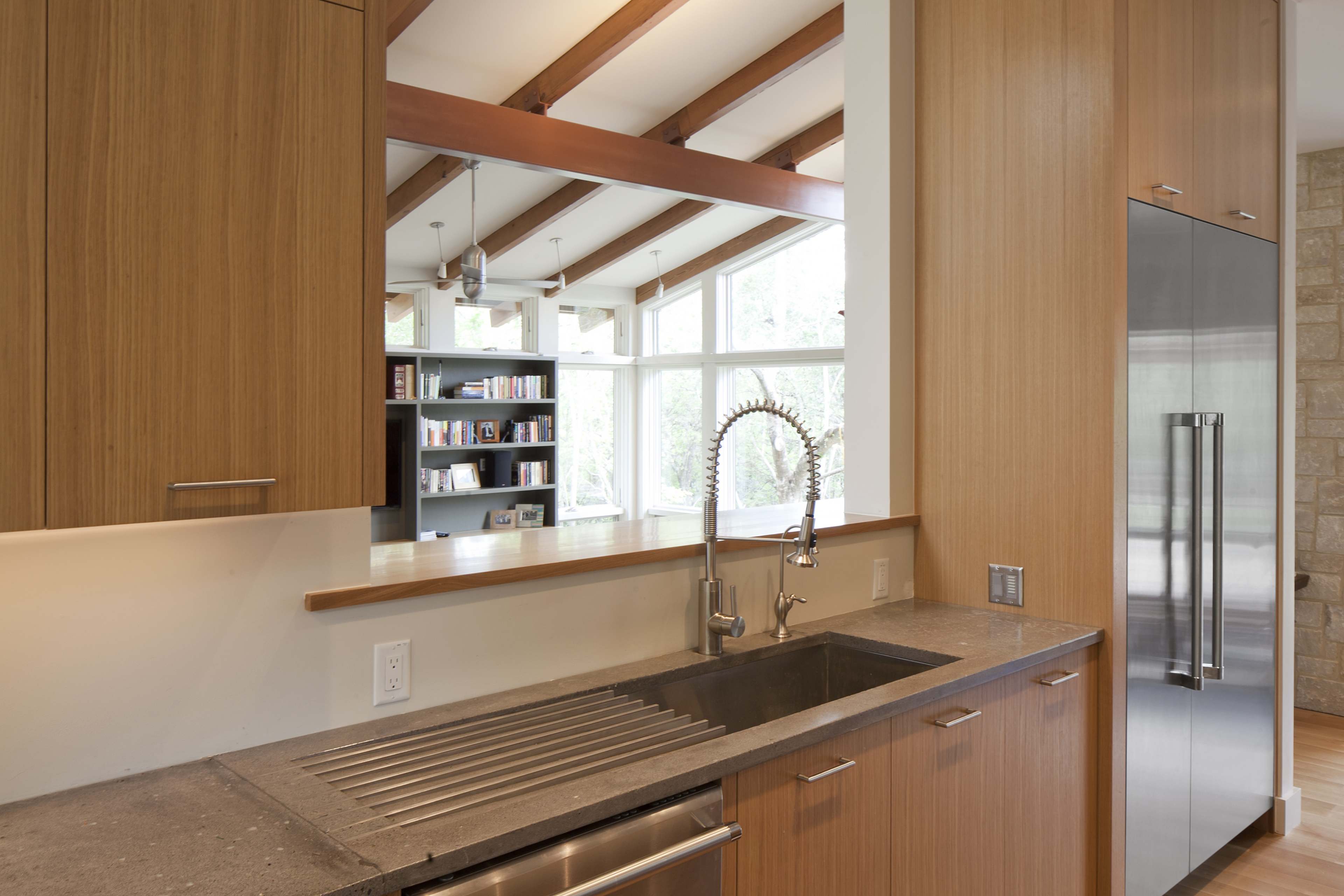 A modern kitchen with wooden cabinetry, a stainless steel refrigerator, and a sink under a large window.