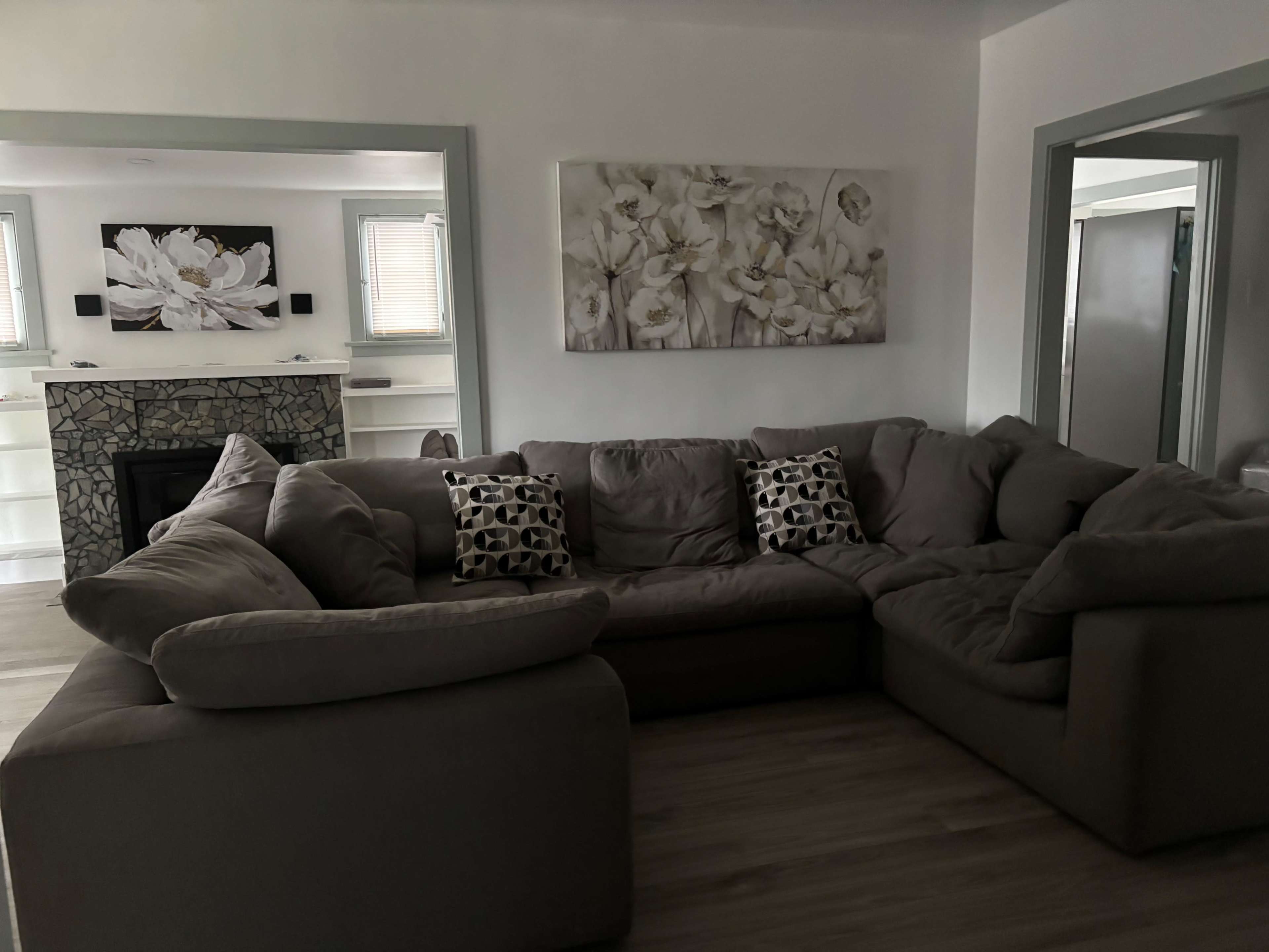 The image depicts a spacious living room with a large gray sectional sofa, two decorative pillows, and a wall featuring floral artwork above a stone fireplace.