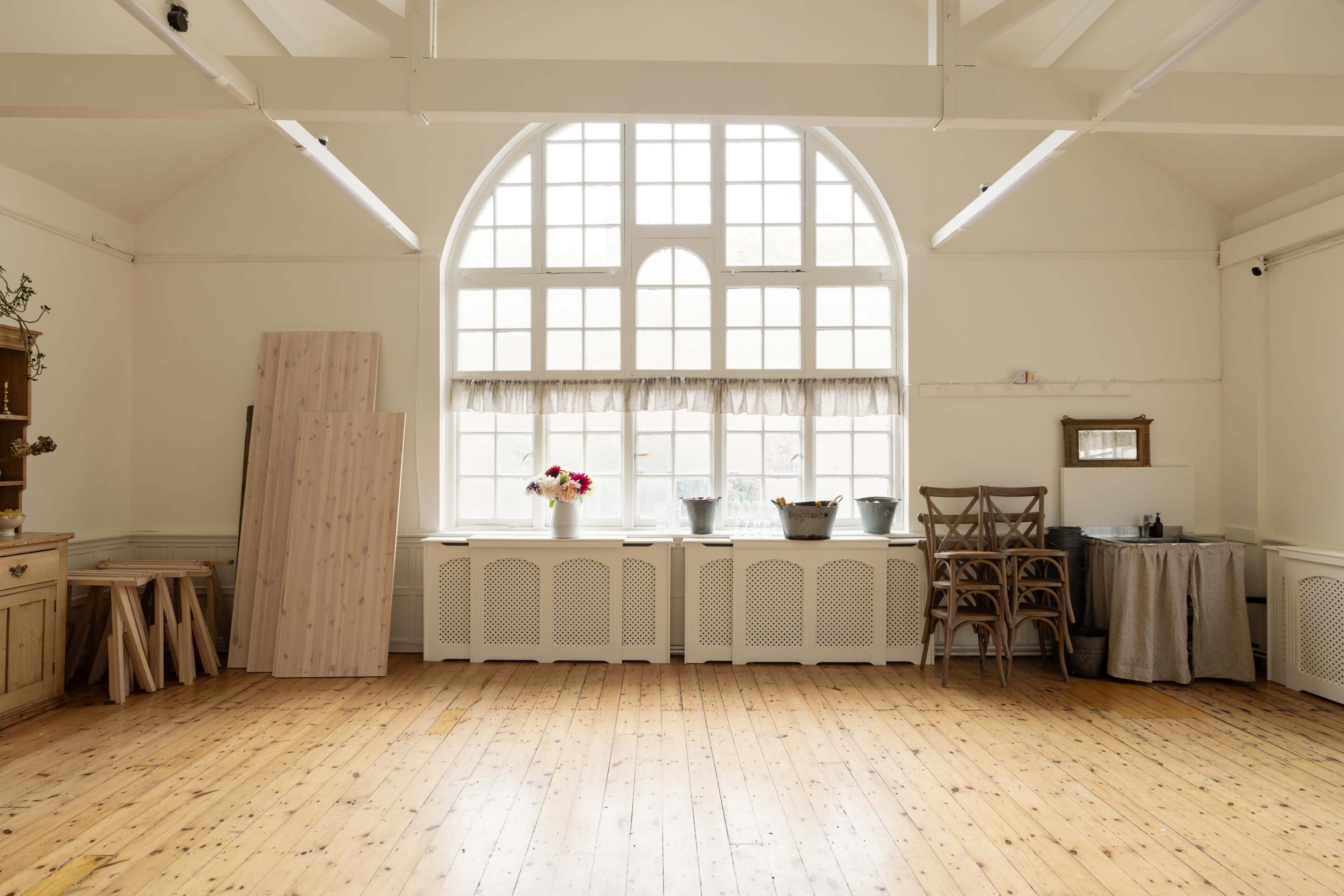 The image shows a spacious room with wooden floors, a large arched window, and various items including planks of wood, a table, and chairs near the walls.