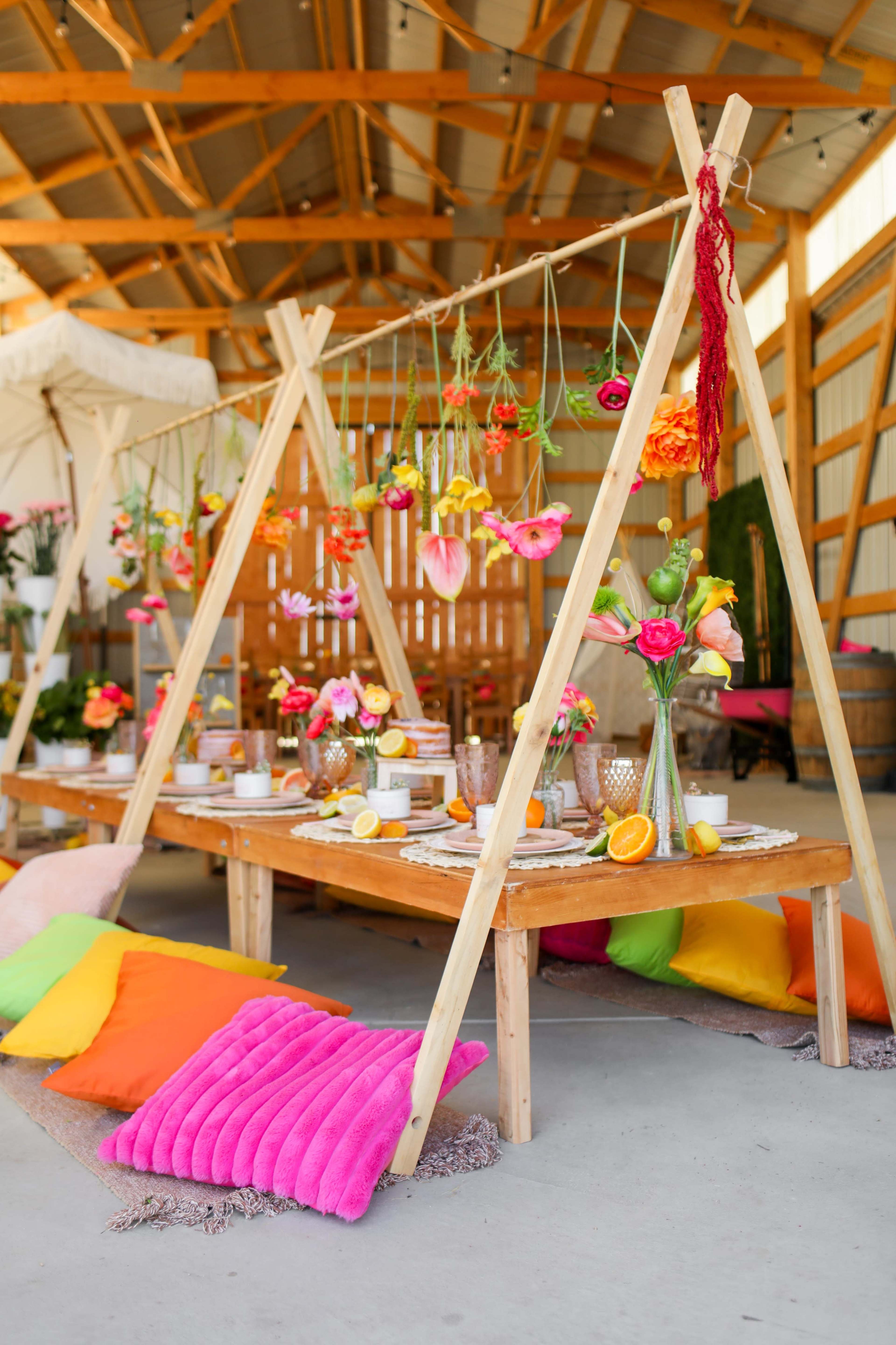 A colorful table setting is arranged under a wooden canopy, featuring vibrant flowers and cushions in a barn-like interior.