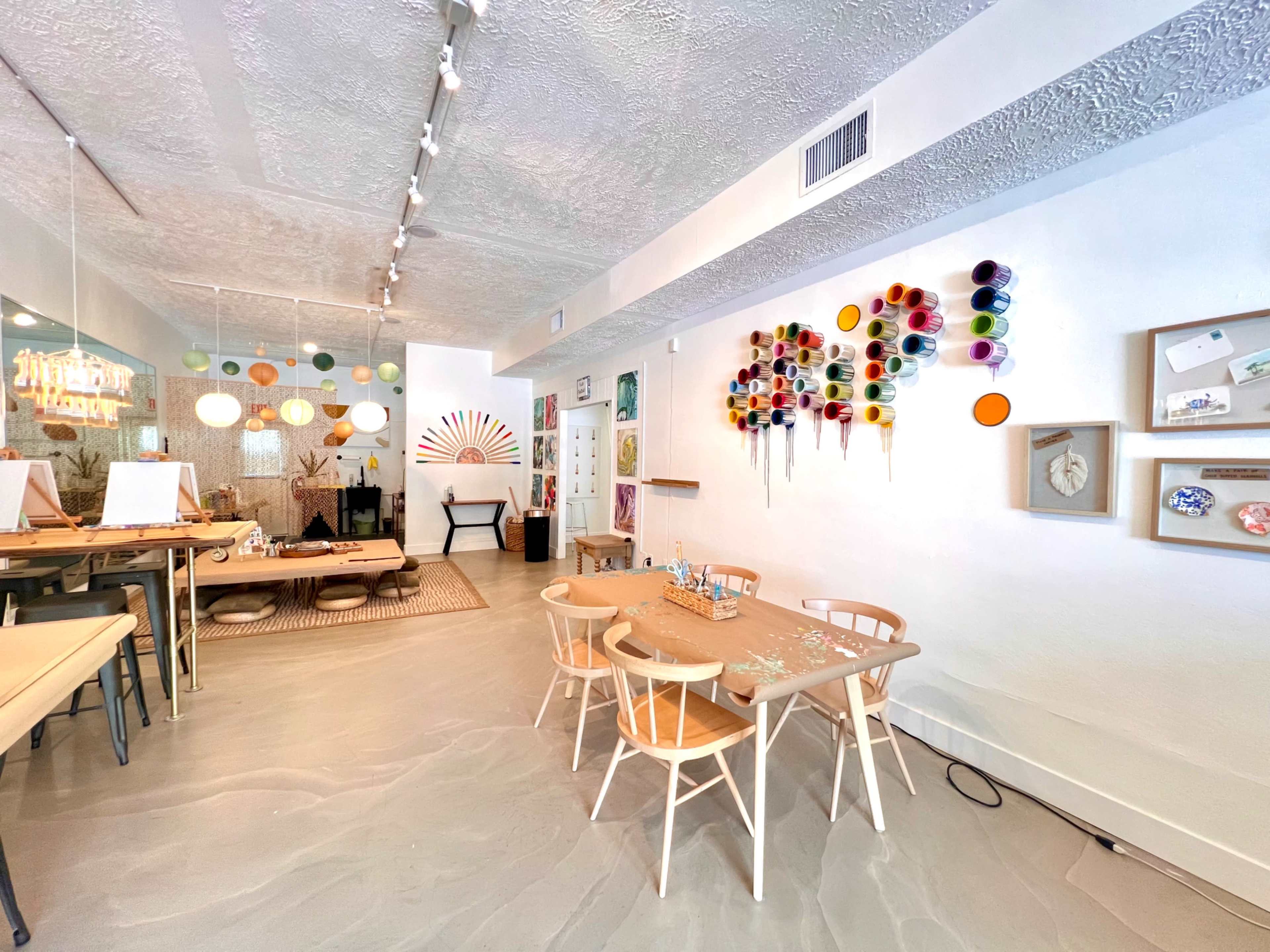 A brightly lit art studio featuring tables with wooden chairs, colorful wall decorations, and an array of art supplies on display.