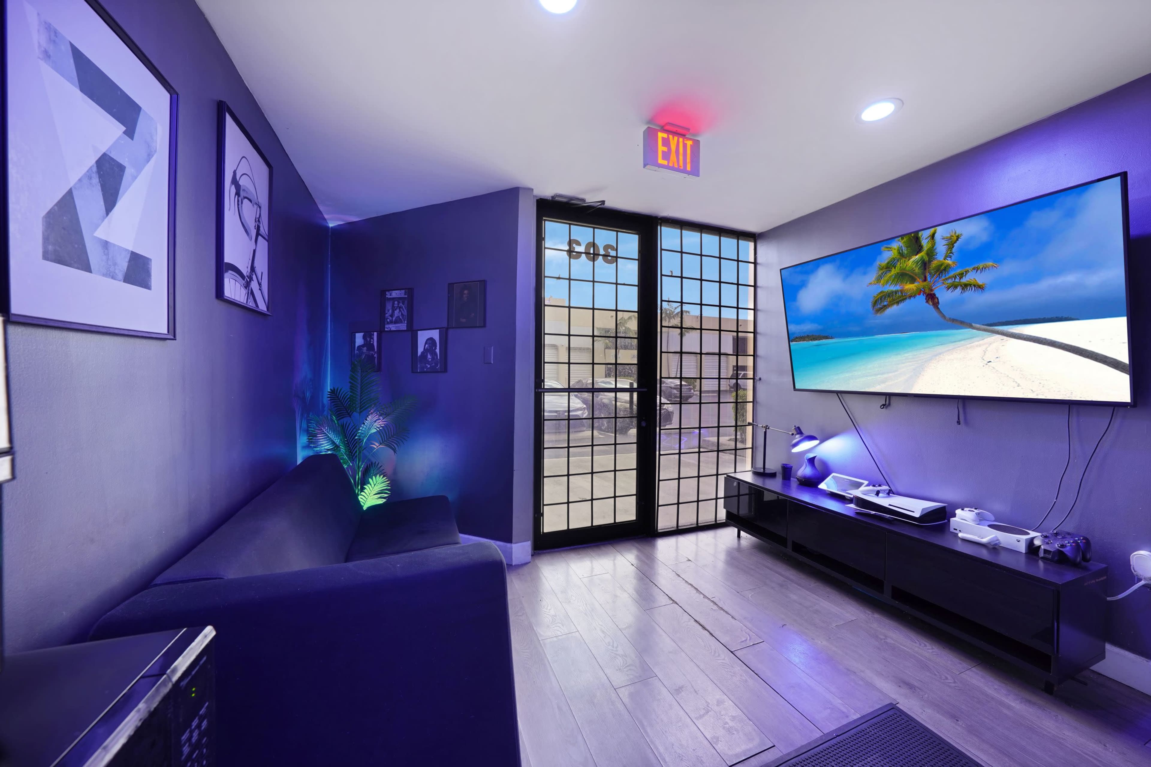 A modern lounge area features a sofa and a large TV displaying a beach scene, with purple lighting and a doorway visible in the background.