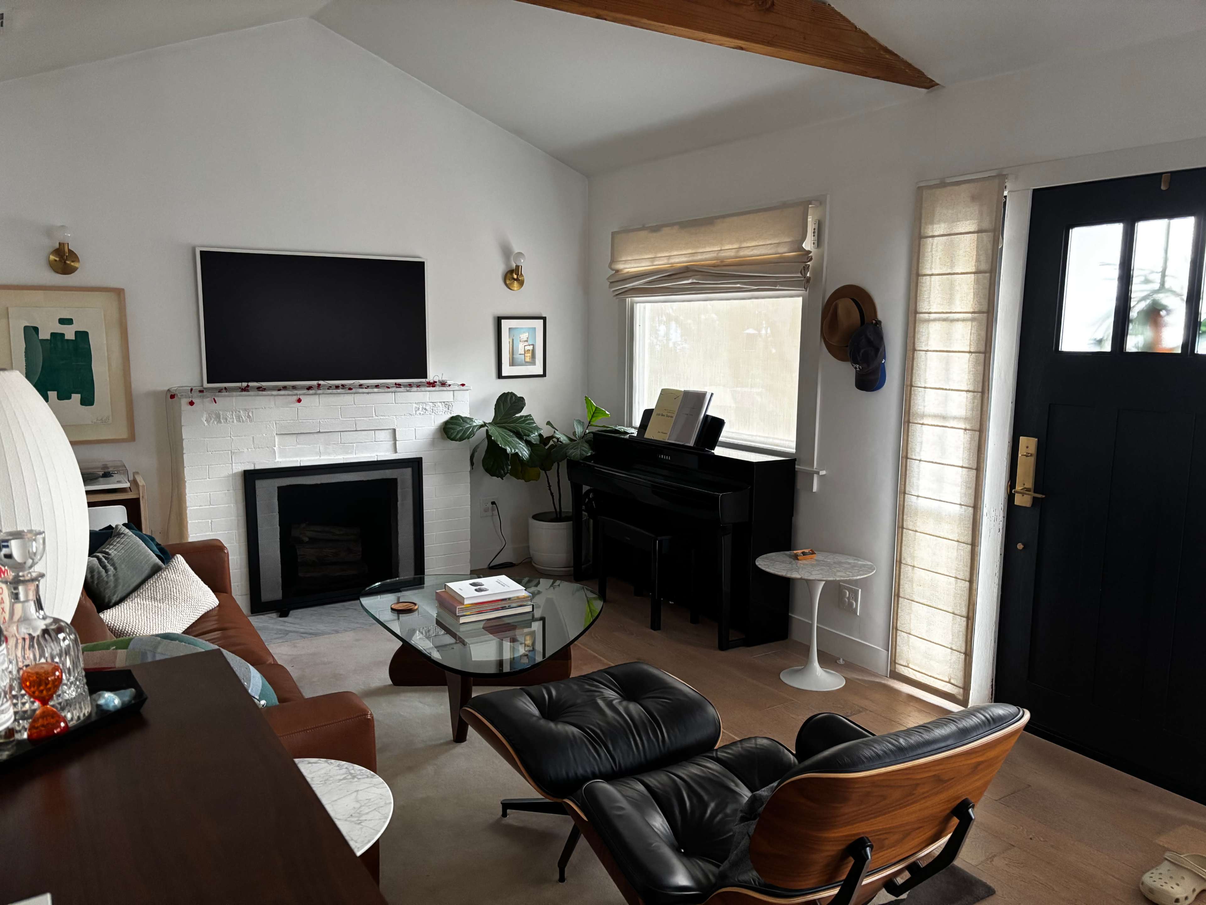 The image shows a living room featuring a couch, a glass coffee table, a piano, and a large television mounted on the wall.