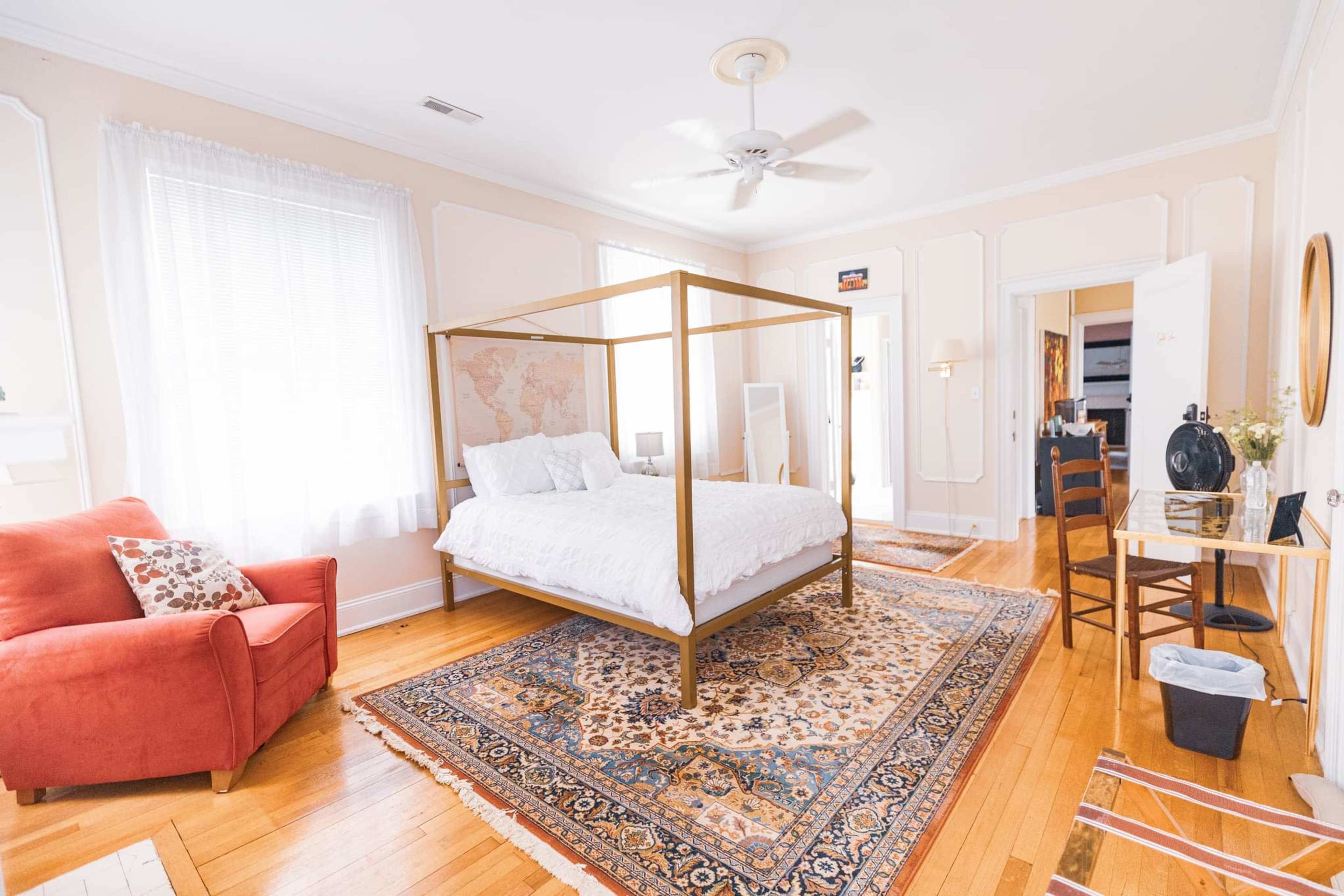 A well-lit bedroom featuring a four-poster bed, a red armchair, and a small wooden desk against a decorative rug.