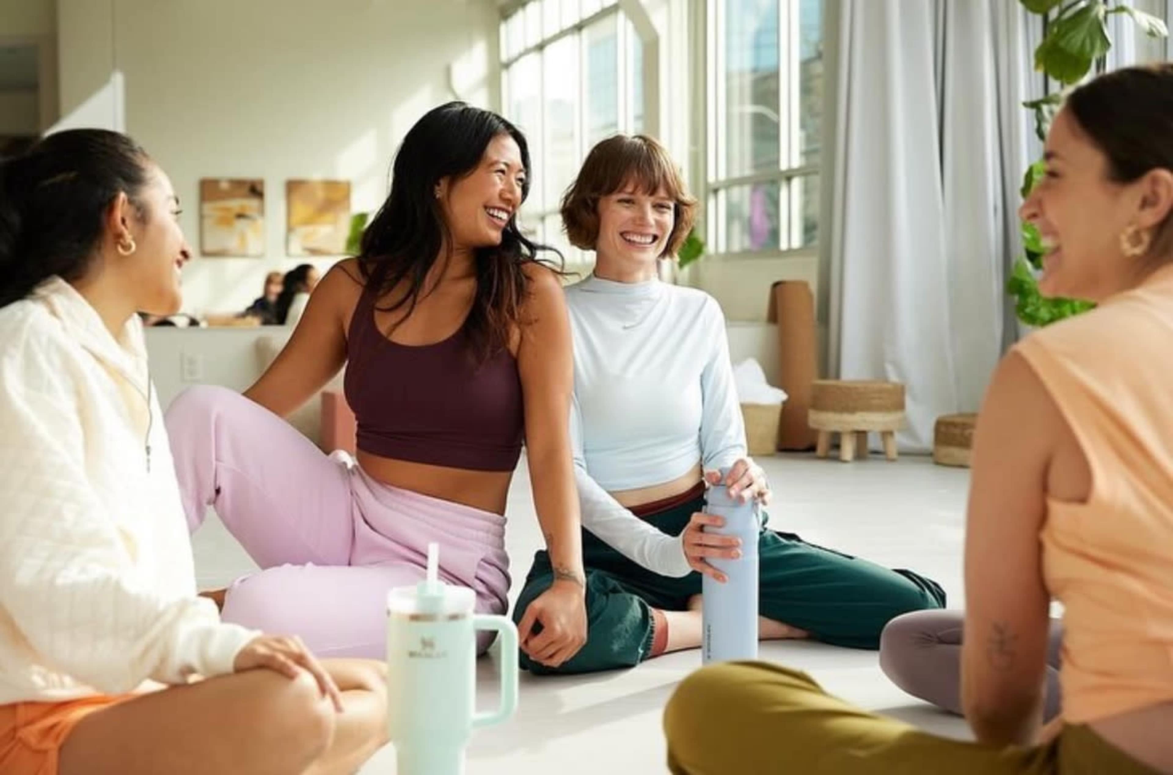 Four women sit together on a bright indoor floor, engaged in conversation while wearing casual athletic clothing.