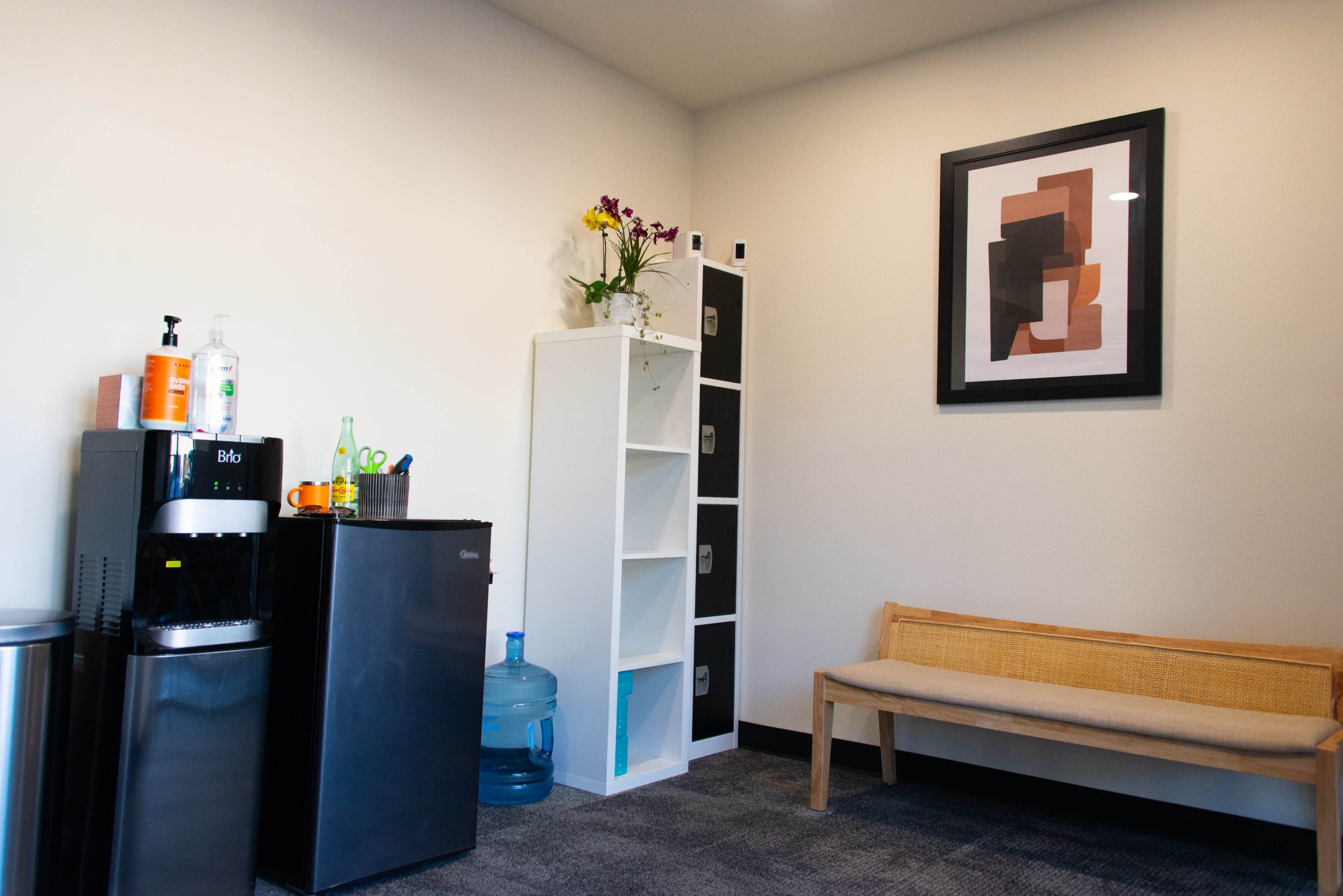 The image shows a small room featuring a black mini-fridge, a white bookshelf with some items, a bench, and a water cooler, along with a framed artwork on the wall.