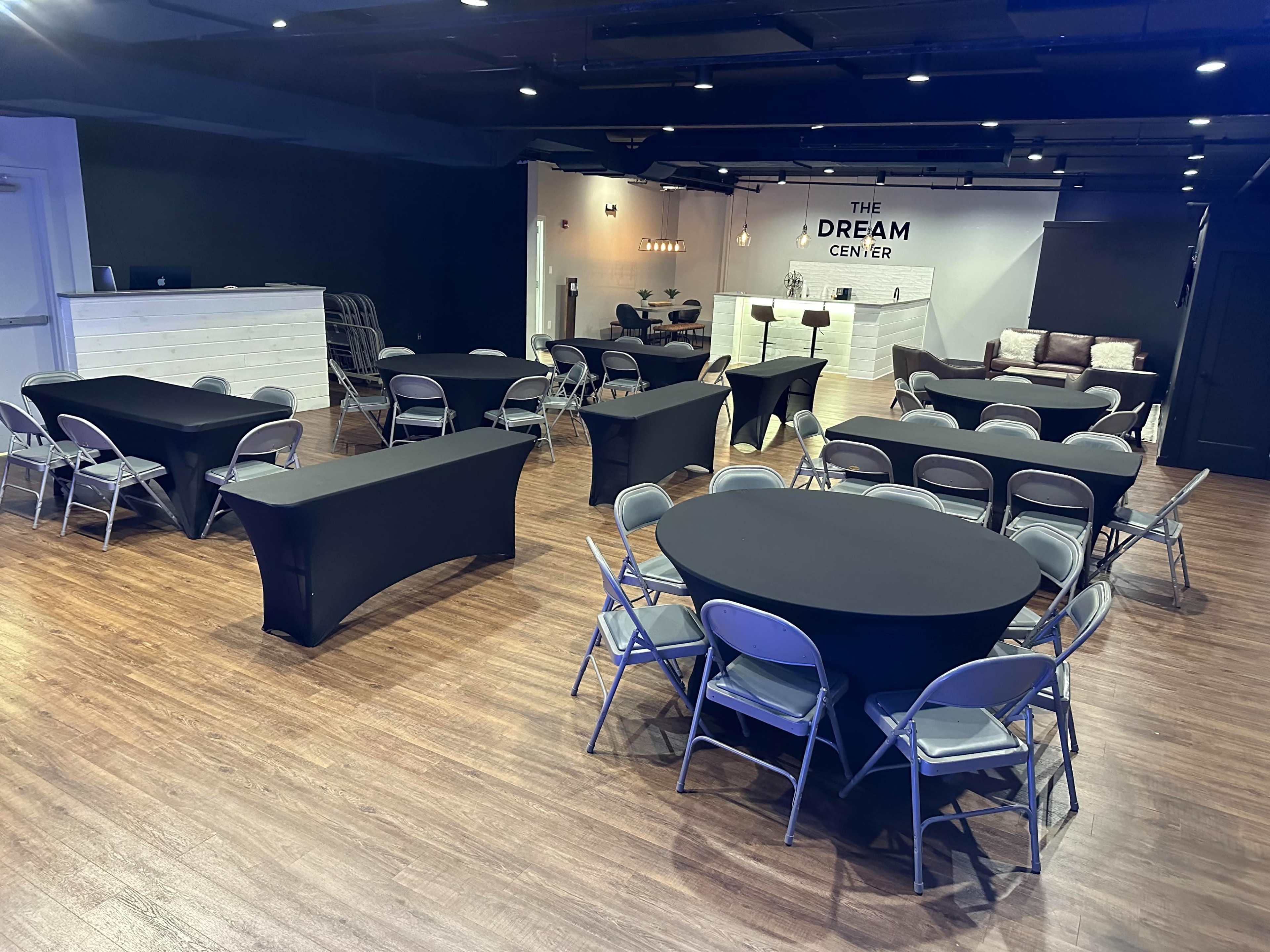 A spacious event room arranged with black table covers and gray folding chairs, featuring a reception area with a sign that reads "THE DREAM CENTER."