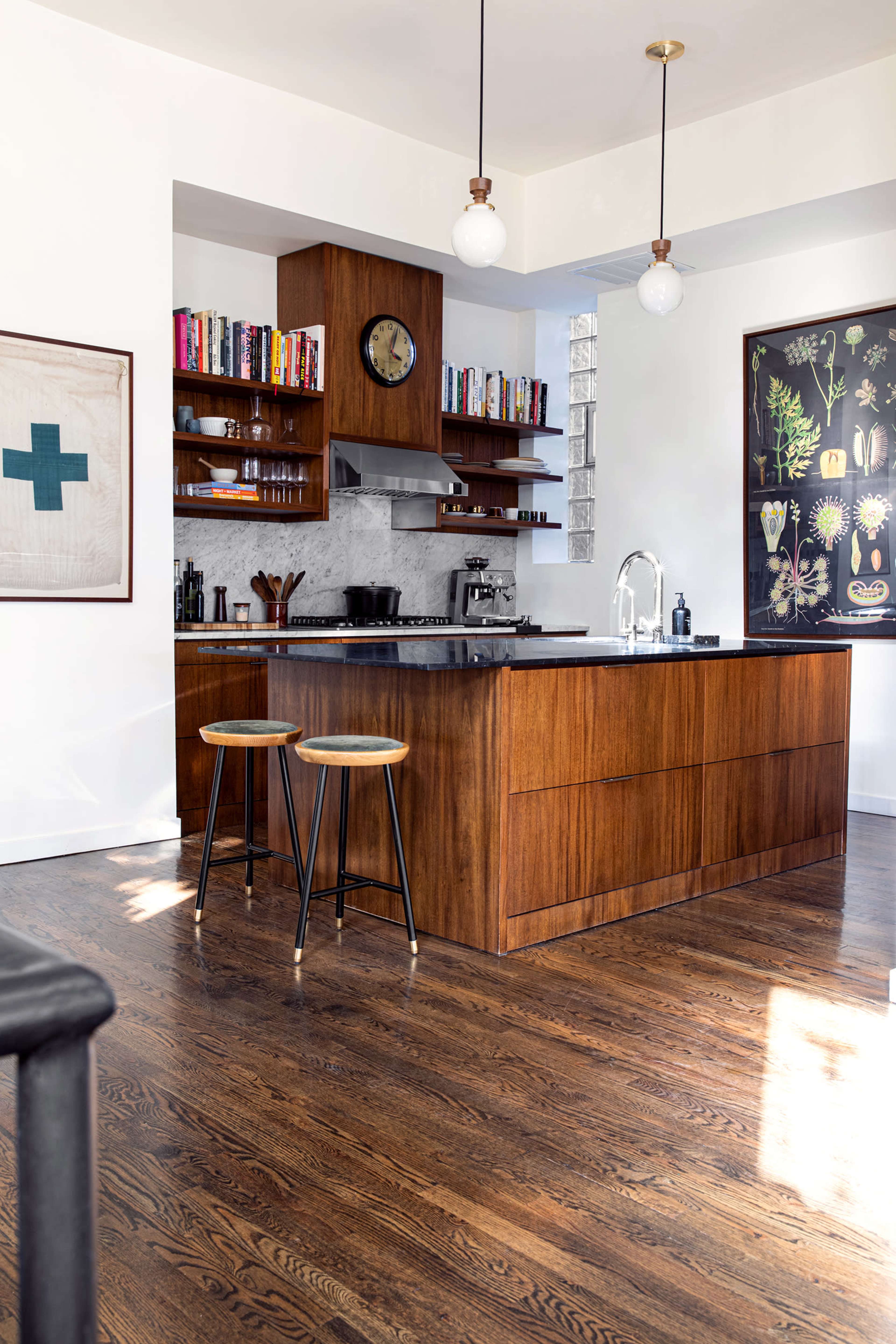 A modern kitchen features a central wooden island with two stools, surrounded by shelves filled with books and kitchen tools, and a wall adorned with decorative artwork.