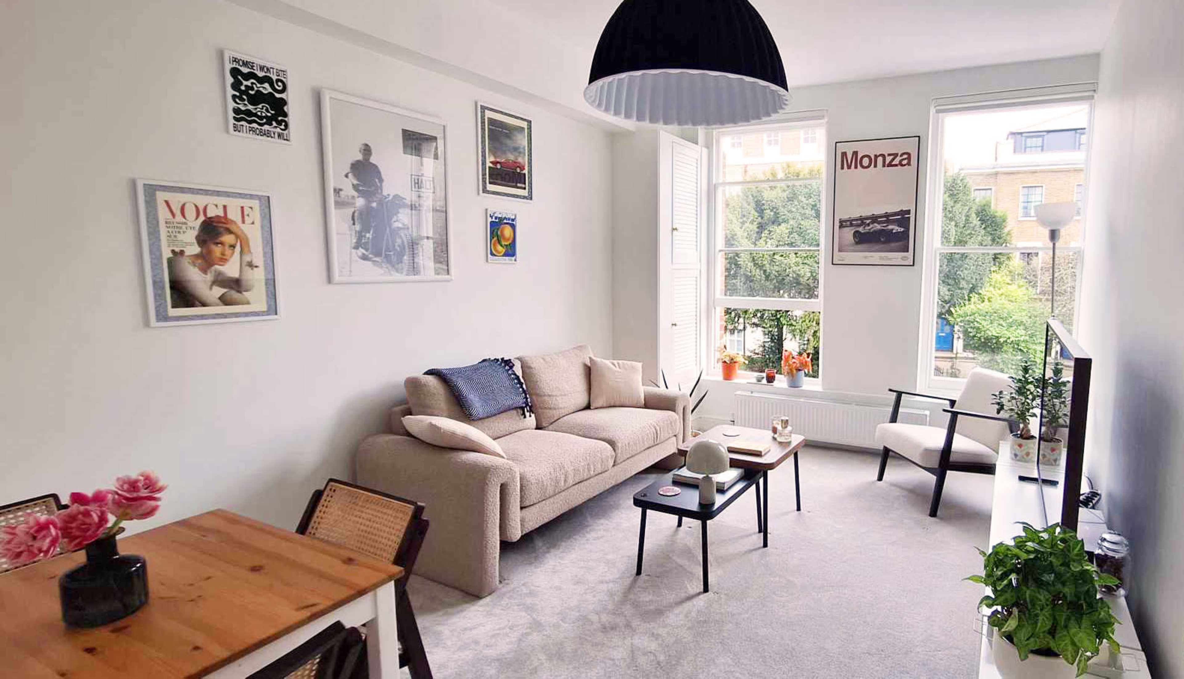 The image shows a cozy living room with a light-colored sofa, a coffee table, a small dining table, and framed artwork on the walls, featuring large windows that let in natural light.