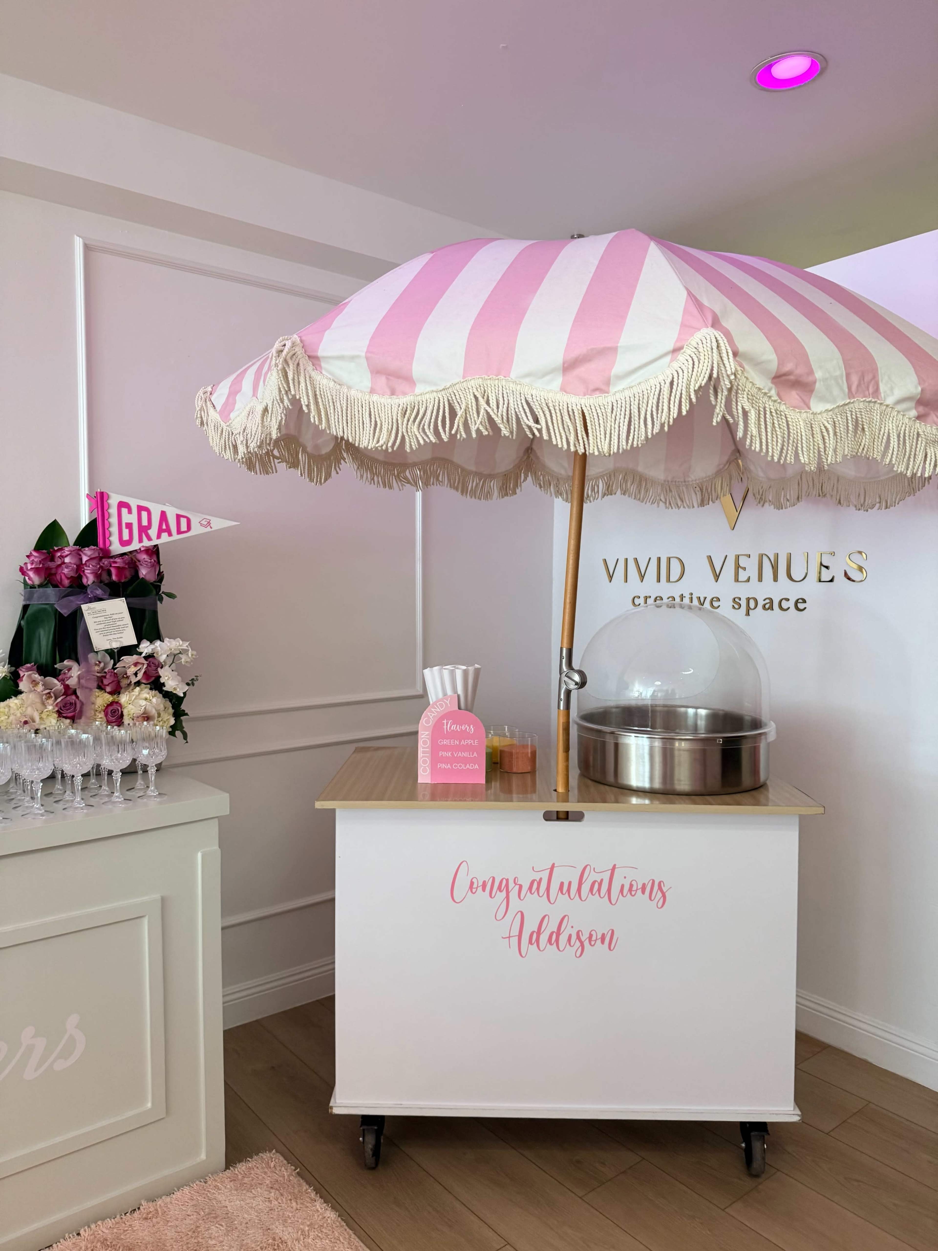 A pink-and-white striped umbrella shades a cart decorated with the words "Congratulations Addison," alongside a floral arrangement and signage in a bright event space.