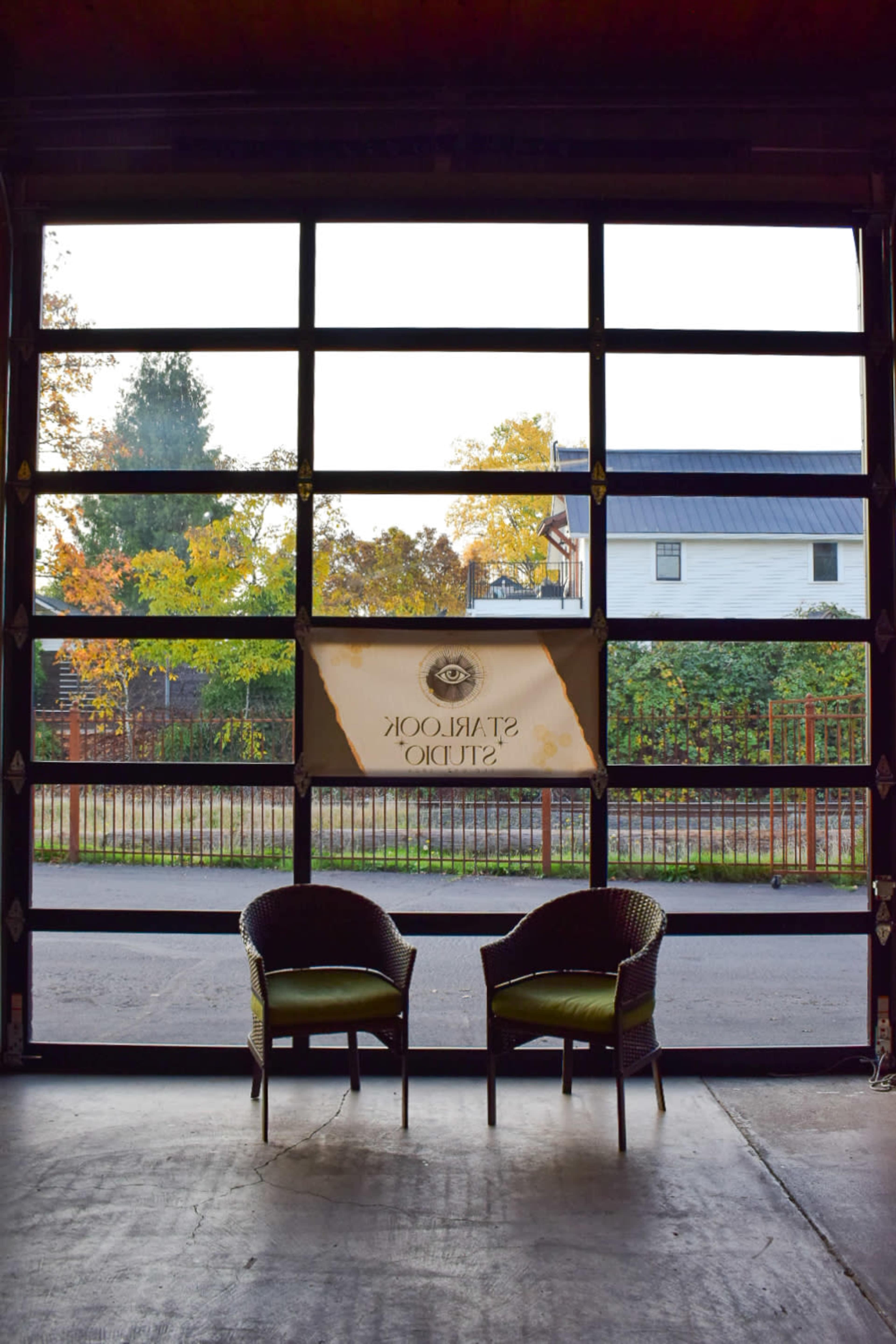Two wicker chairs are positioned inside an industrial-style garage, facing an open door that reveals a view of trees and a white house across the street.
