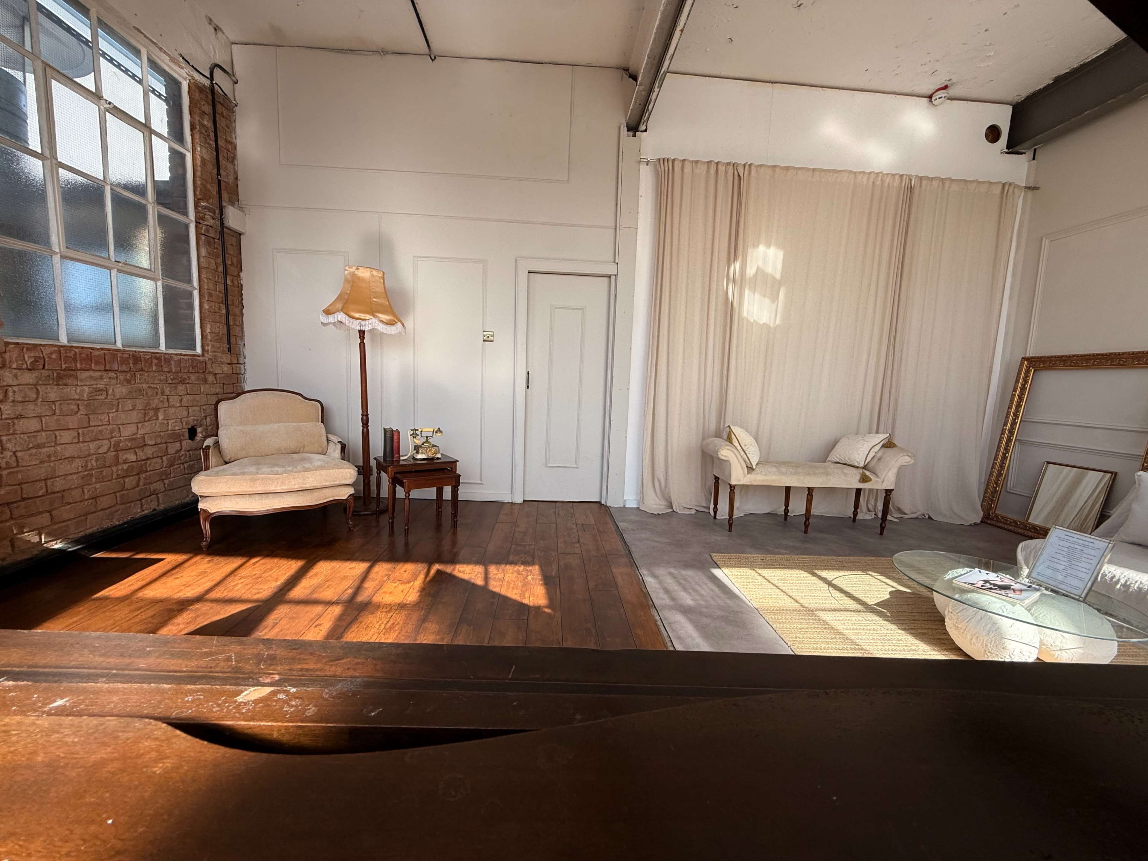 The image shows a spacious room with wooden flooring, featuring two upholstered chairs, a small table, and a large window that lets in natural light.
